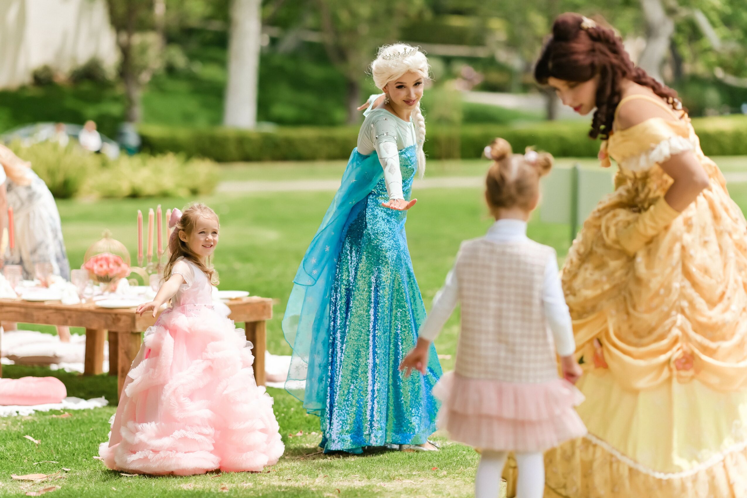 Children and a woman in princess costumes outdoors, with a picnic setup and greenery in the background.