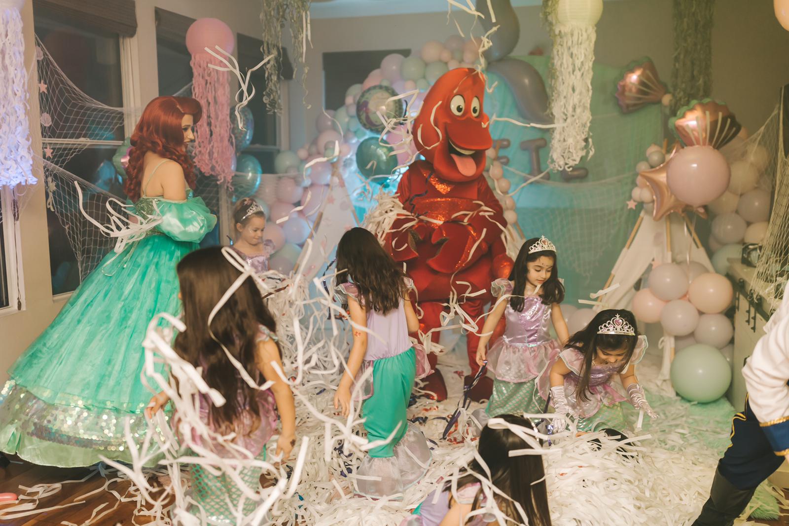 Children and a person in a red dragon costume at a party with balloons and decorations, some children wearing princess costumes.