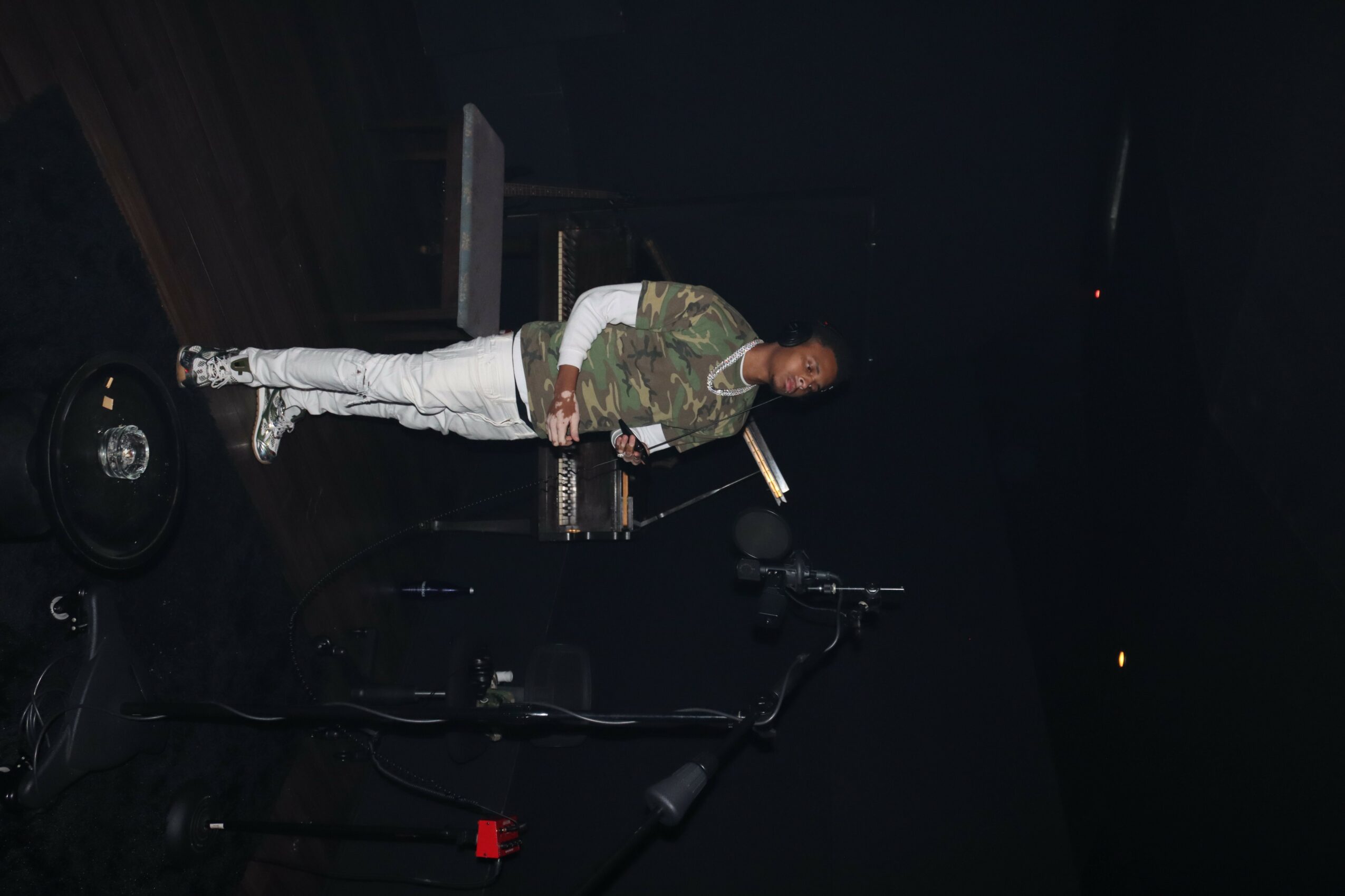 Person standing in a dark room, wearing camouflage jacket and white pants, with equipment on the floor.