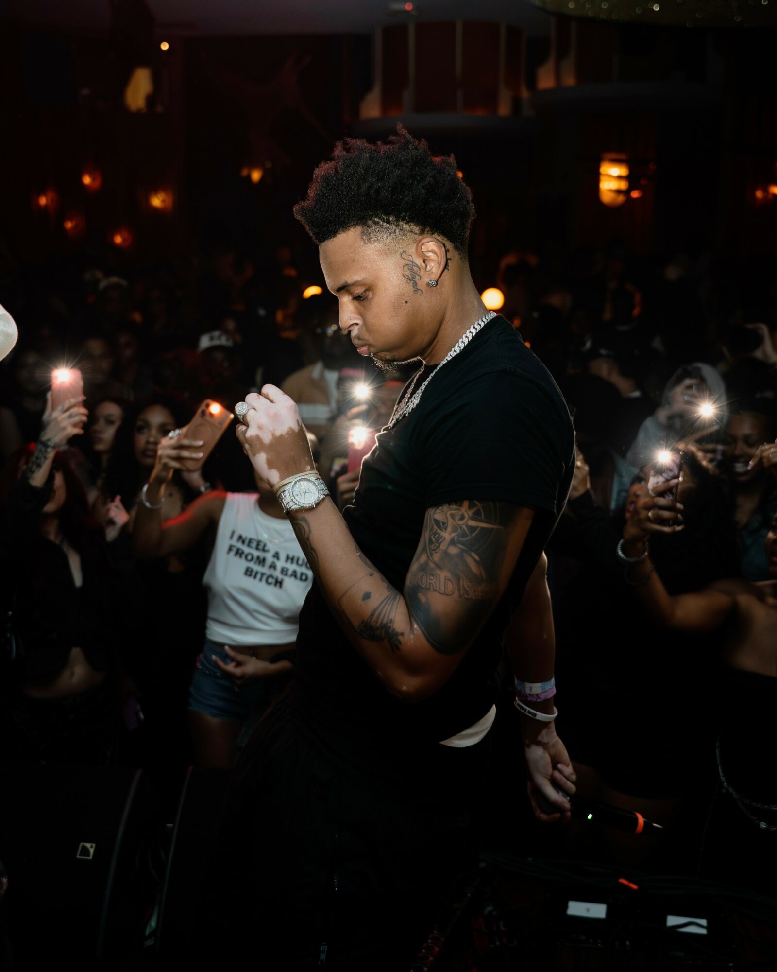 Young man with tattoos and jewelry looking down in a dimly lit crowd with phone flashes around him.