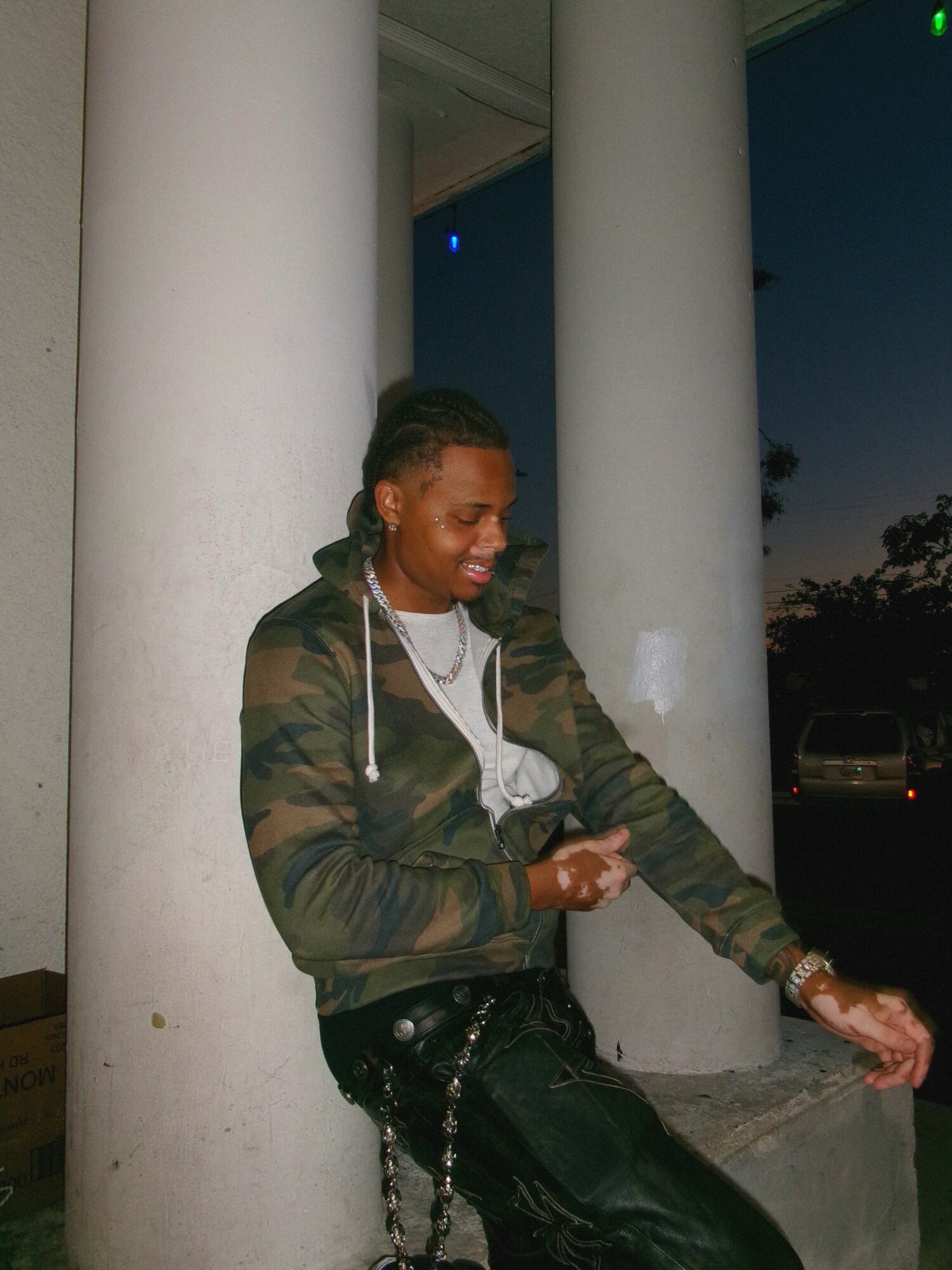 Person standing outdoors near large white columns, smiling, wearing a camouflage jacket and black pants, during evening or night.