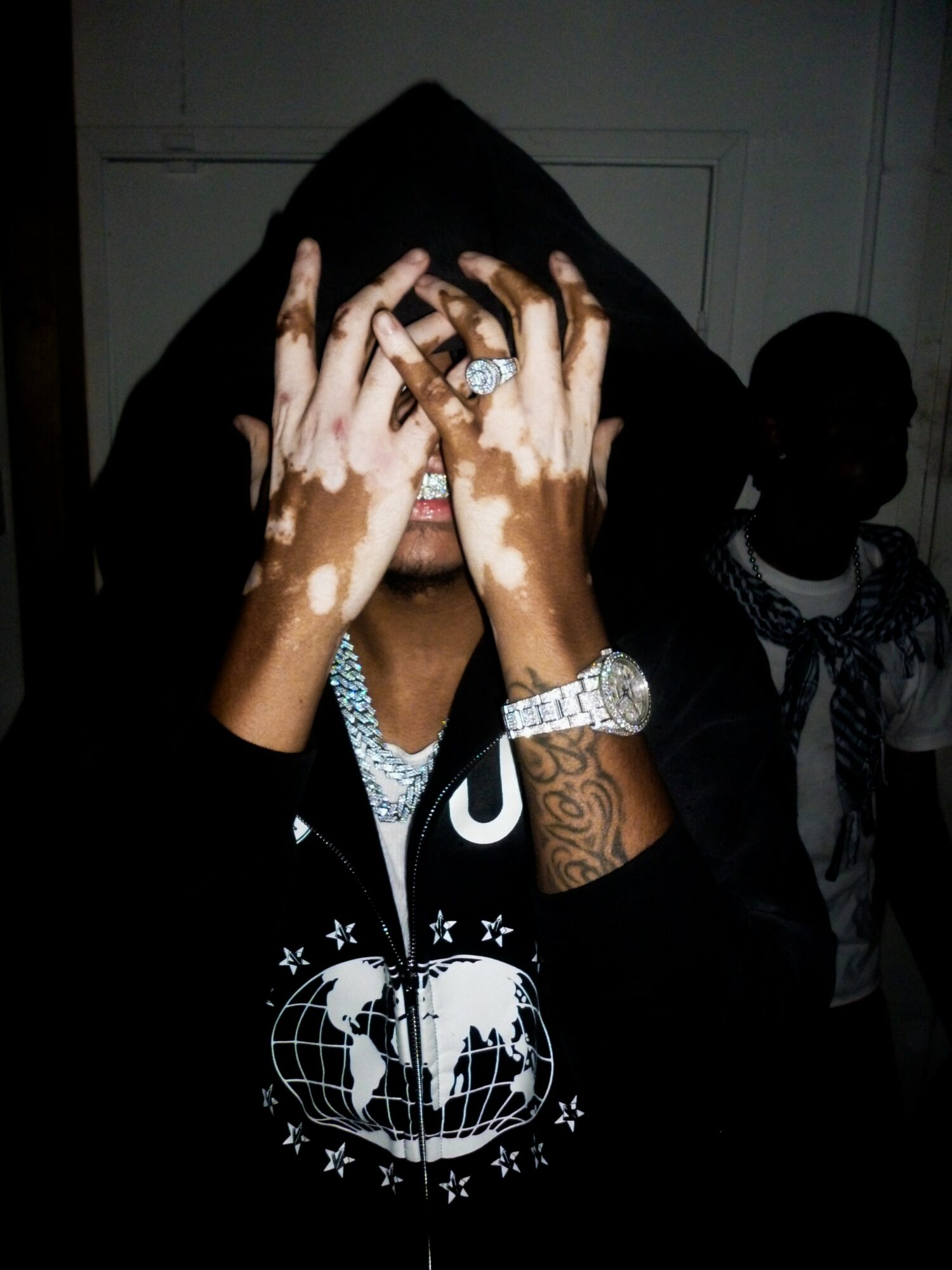 Person with hands covering face, wearing a black hoodie, jewelry, and a black jacket with a globe design, in a dark setting.