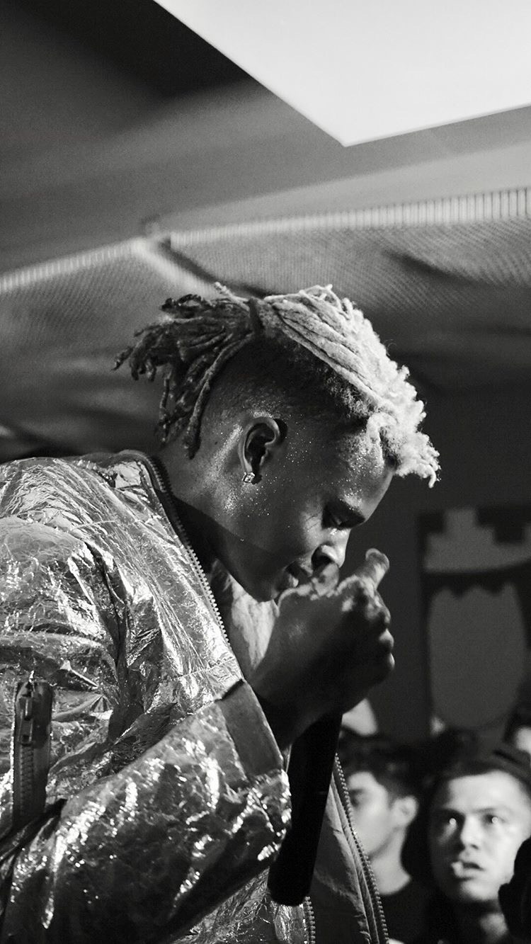 Person with styled hair and patterned jacket holding a microphone, with a group of people in the background.