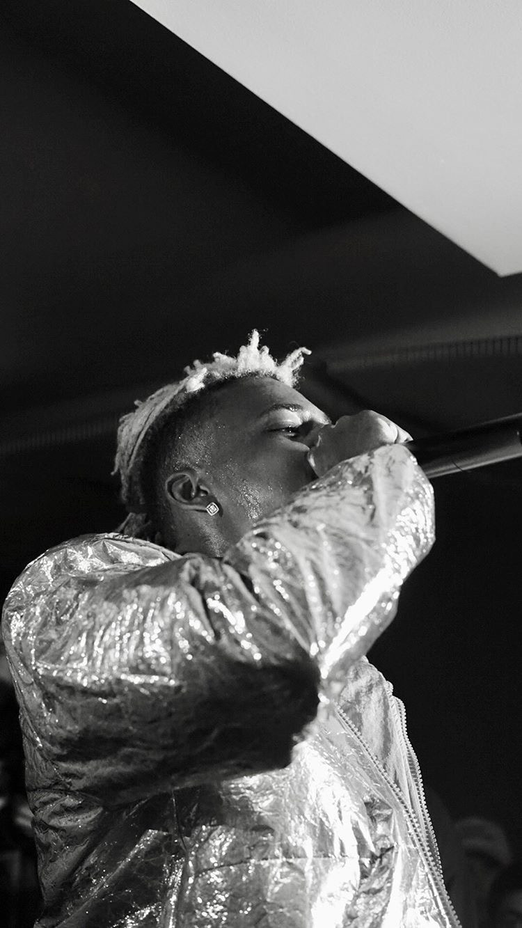 Person with short hair and shiny jacket, covering their face with their hand, in a dark indoor setting.