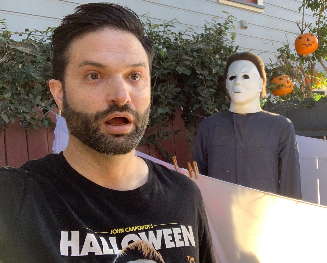 A man with dark hair and beard takes a selfie outdoors, with a Halloween decoration of a person in a mask and pumpkins in the background.