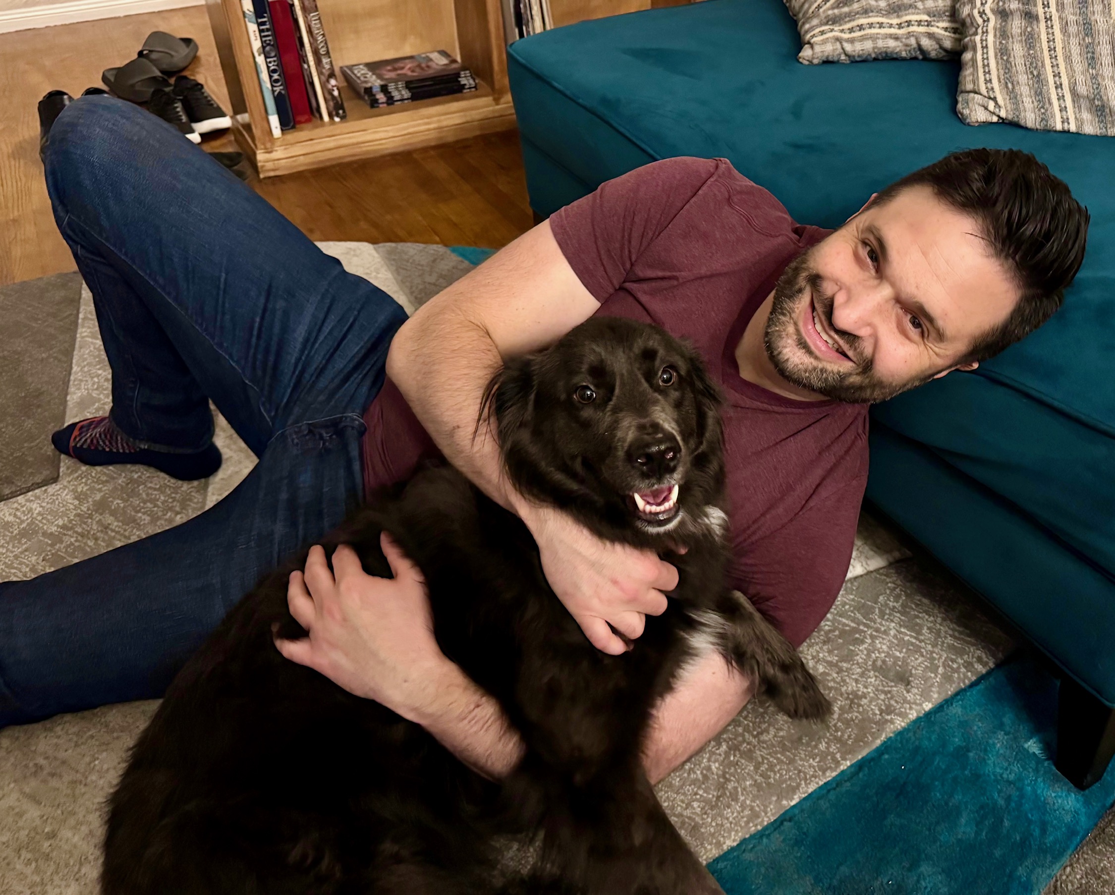 Man lying on the floor hugging a black dog, smiling, in a living room with a blue sofa and bookshelf.