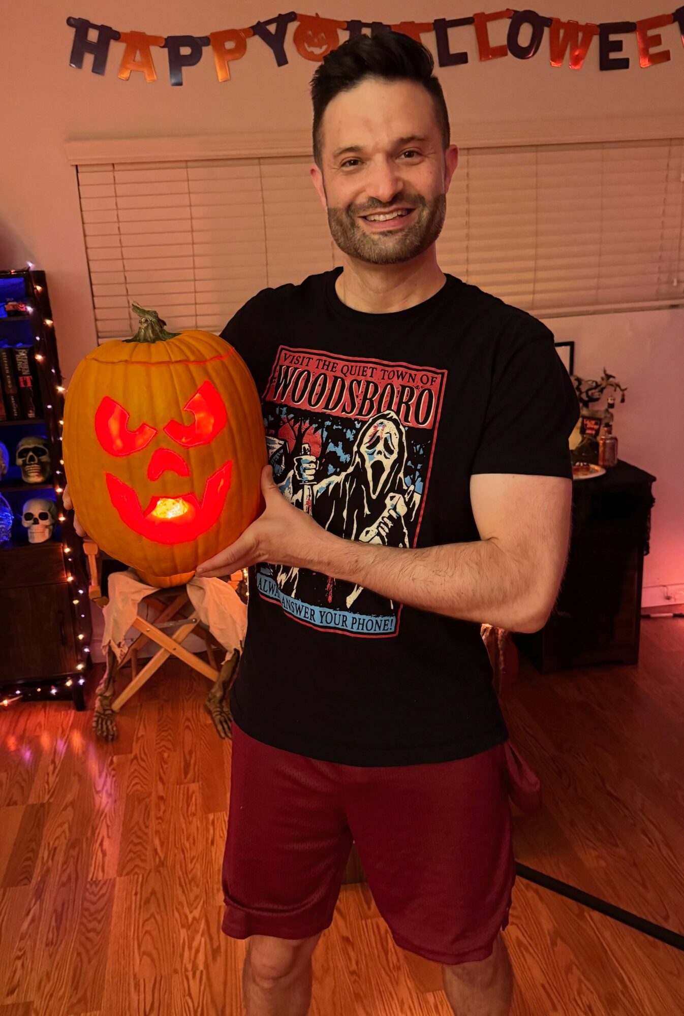 Man smiling holding a carved pumpkin with a glowing face, Halloween decorations in background, festive atmosphere.