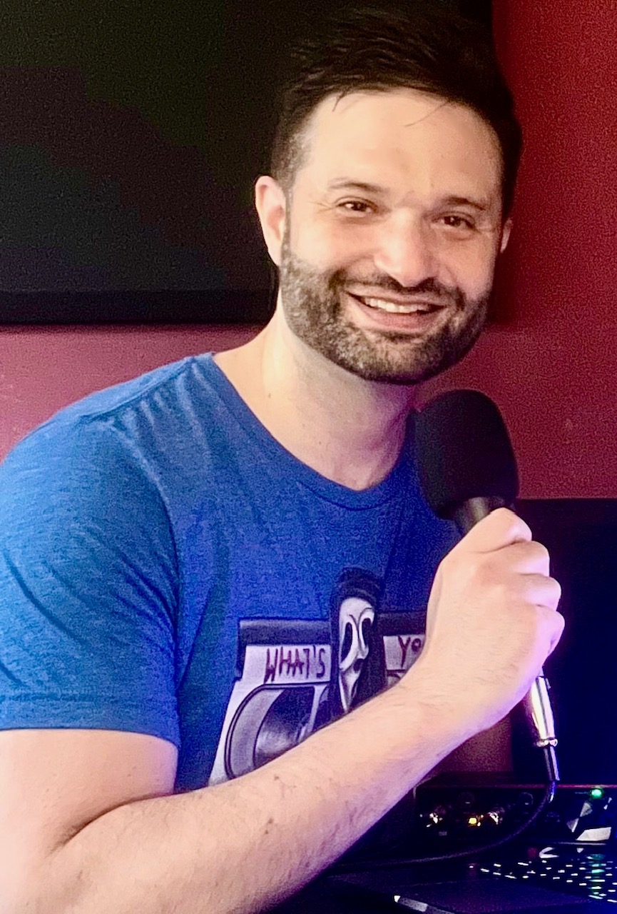 Man with dark hair and beard smiling, holding a microphone, wearing a blue T-shirt, in front of a dark background.