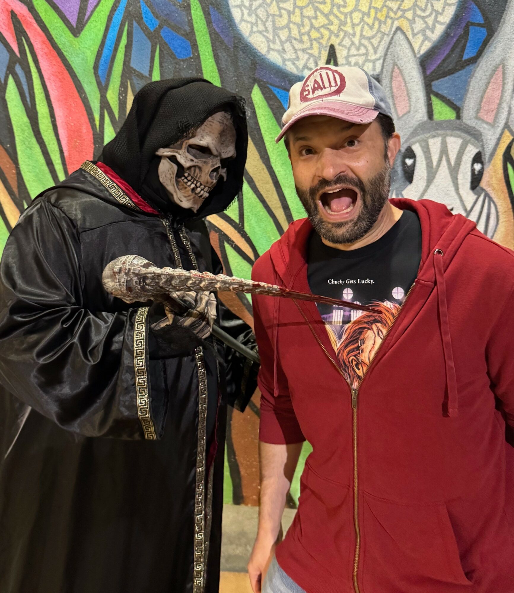 Person in a skeleton costume with a hood and a man in a red hoodie smiling, in front of colorful graffiti art.