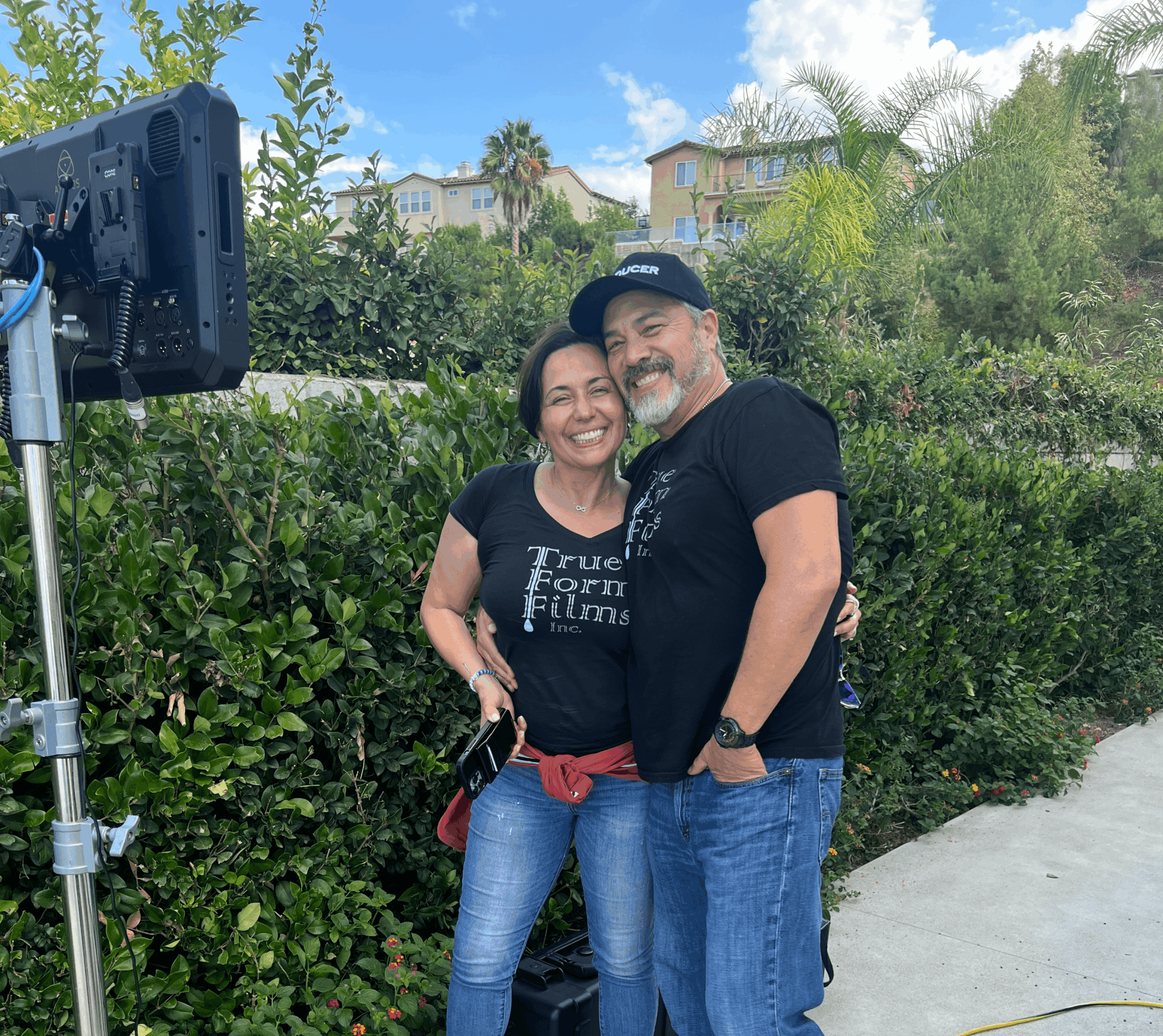 Two people smiling outdoors with greenery and houses in background, standing close together, one with arm around the other.
