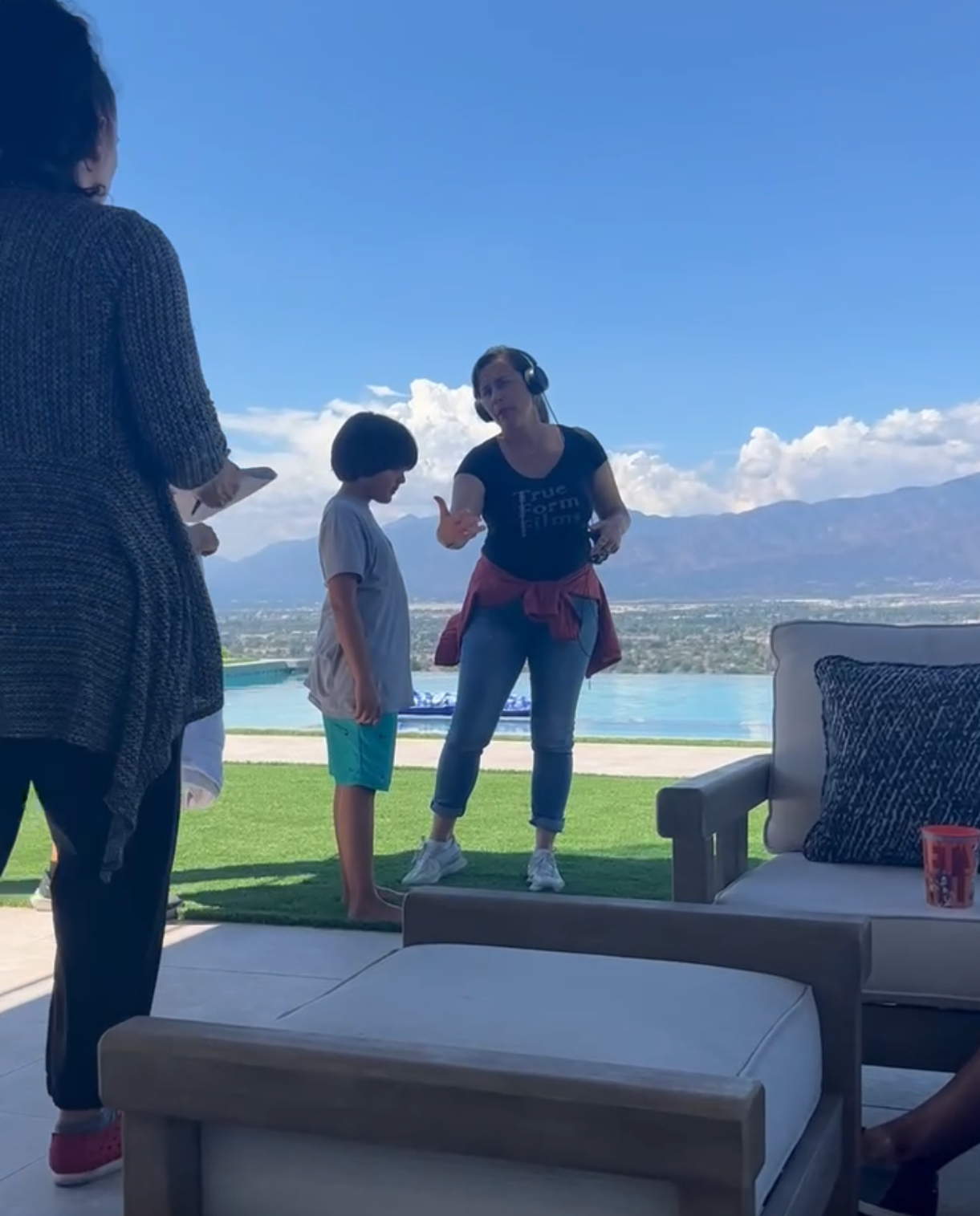 Three people outdoors on a patio with mountains and water in the background, two women and a child, engaging in conversation.
