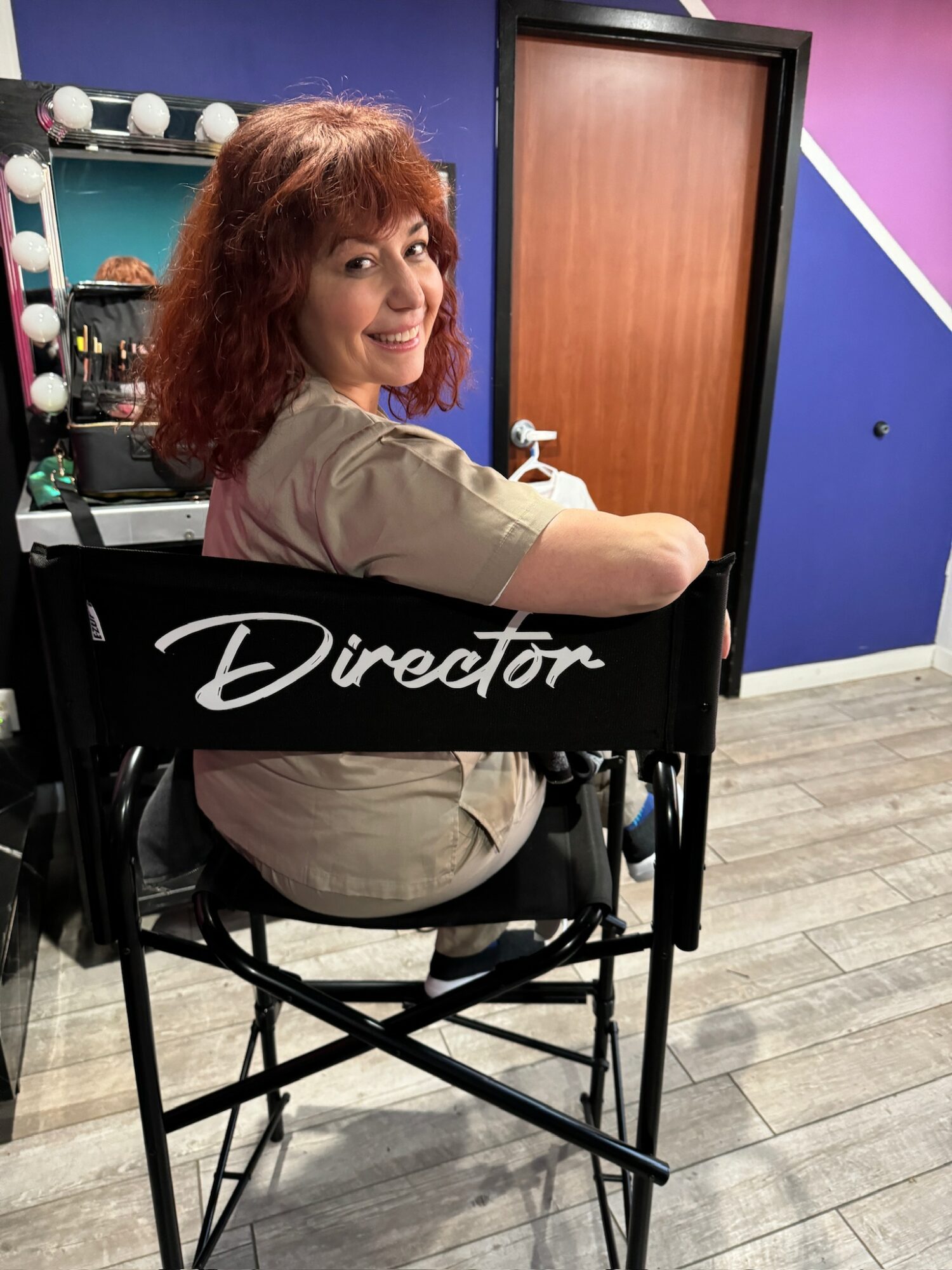 Woman with curly hair sitting in a director's chair, smiling, in a colorful room with a door and makeup station.