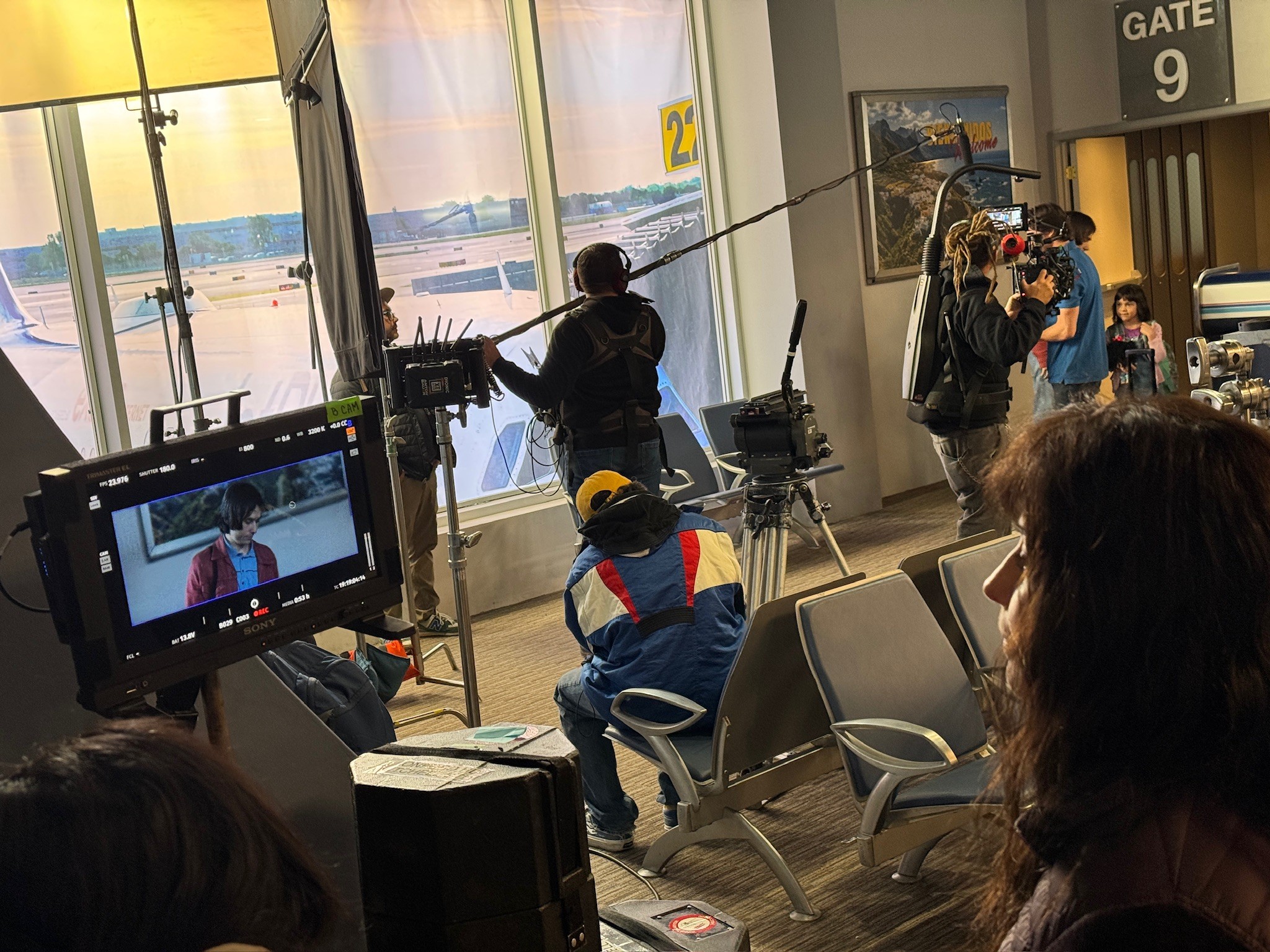 Person fishing through large airport window, crew filming nearby, seated and standing people, airport gate 9 sign, scenic view outside.