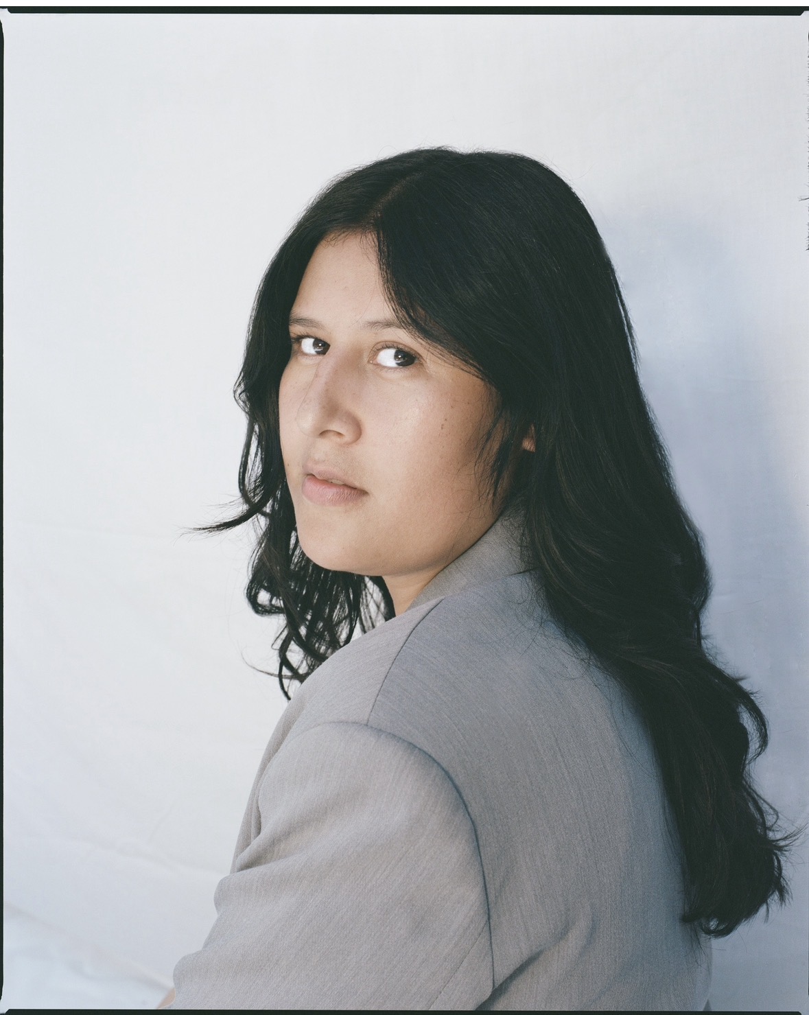 Woman with long dark hair wearing a light gray blazer, looking over her shoulder at the camera.