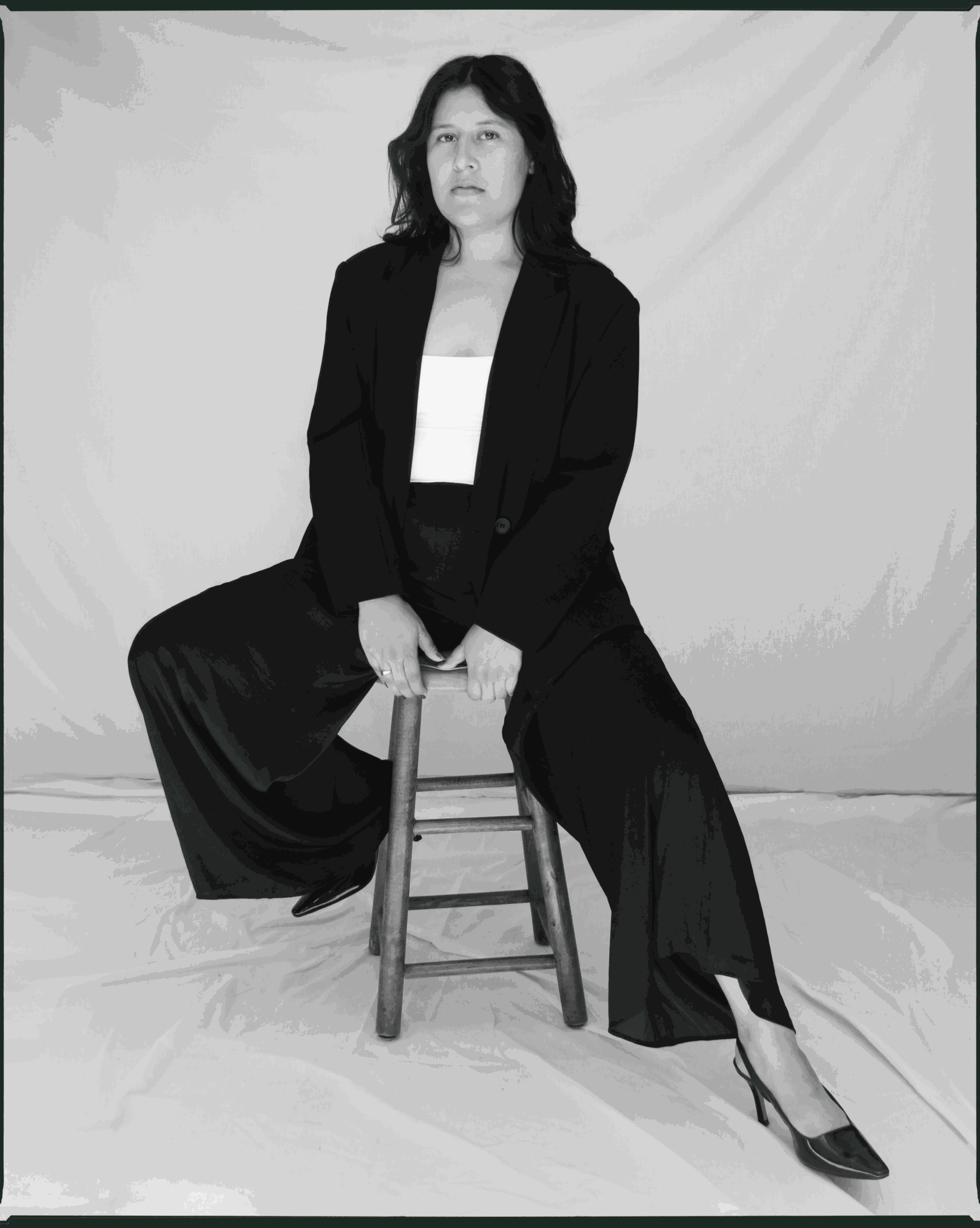 Woman sitting on a stool, wearing a black blazer and wide-leg pants, with a white top, in front of a plain backdrop.