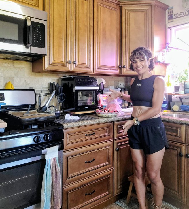 Woman in black outfit standing in kitchen, smiling, with a cake on the counter, wooden cabinets, microwave, and window with sunlight.