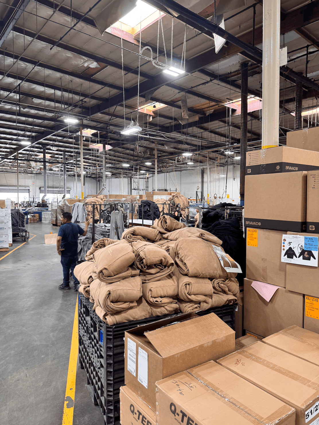 Warehouse interior with stacked bags and boxes, people working, high ceiling with lights and metal beams.