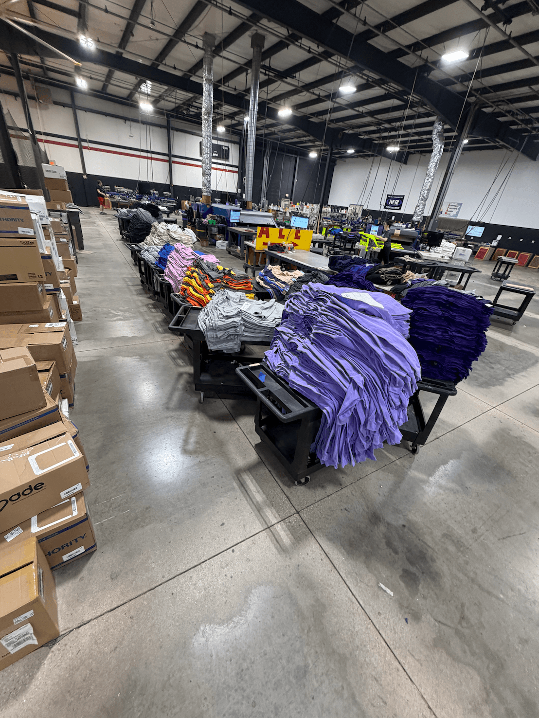Warehouse interior with tables of folded clothing, boxes on the floor, and industrial ceiling with lighting.