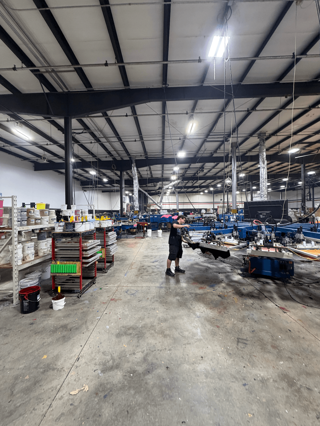 Person standing in large warehouse with shelves, equipment, and machinery, high ceiling with lights, concrete floor, and industrial setting.