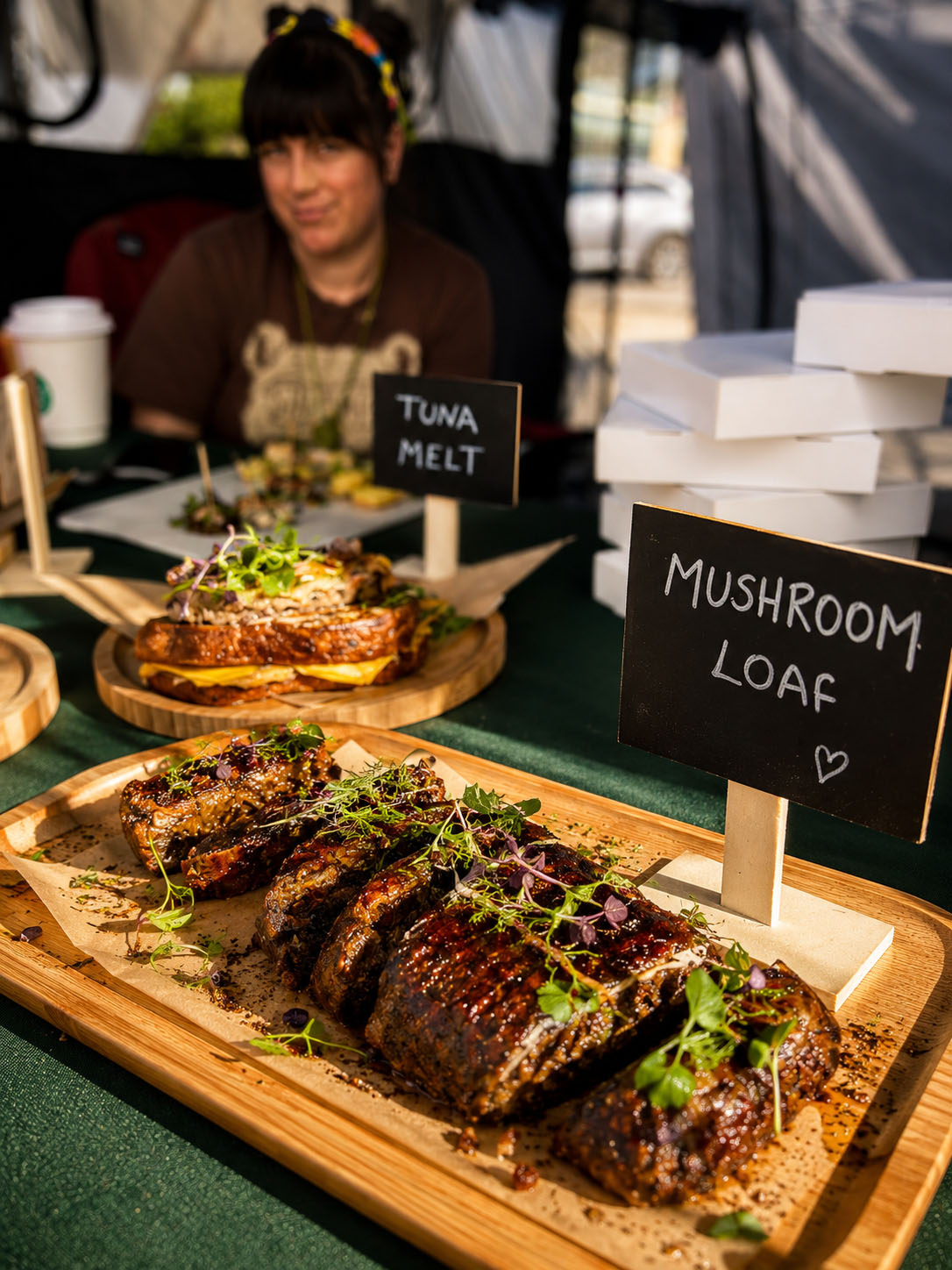 Display of mushroom loaf and tuna melt sandwiches at a food stall, with a woman in the background. The mushroom loaf is sliced and garnished with greens.