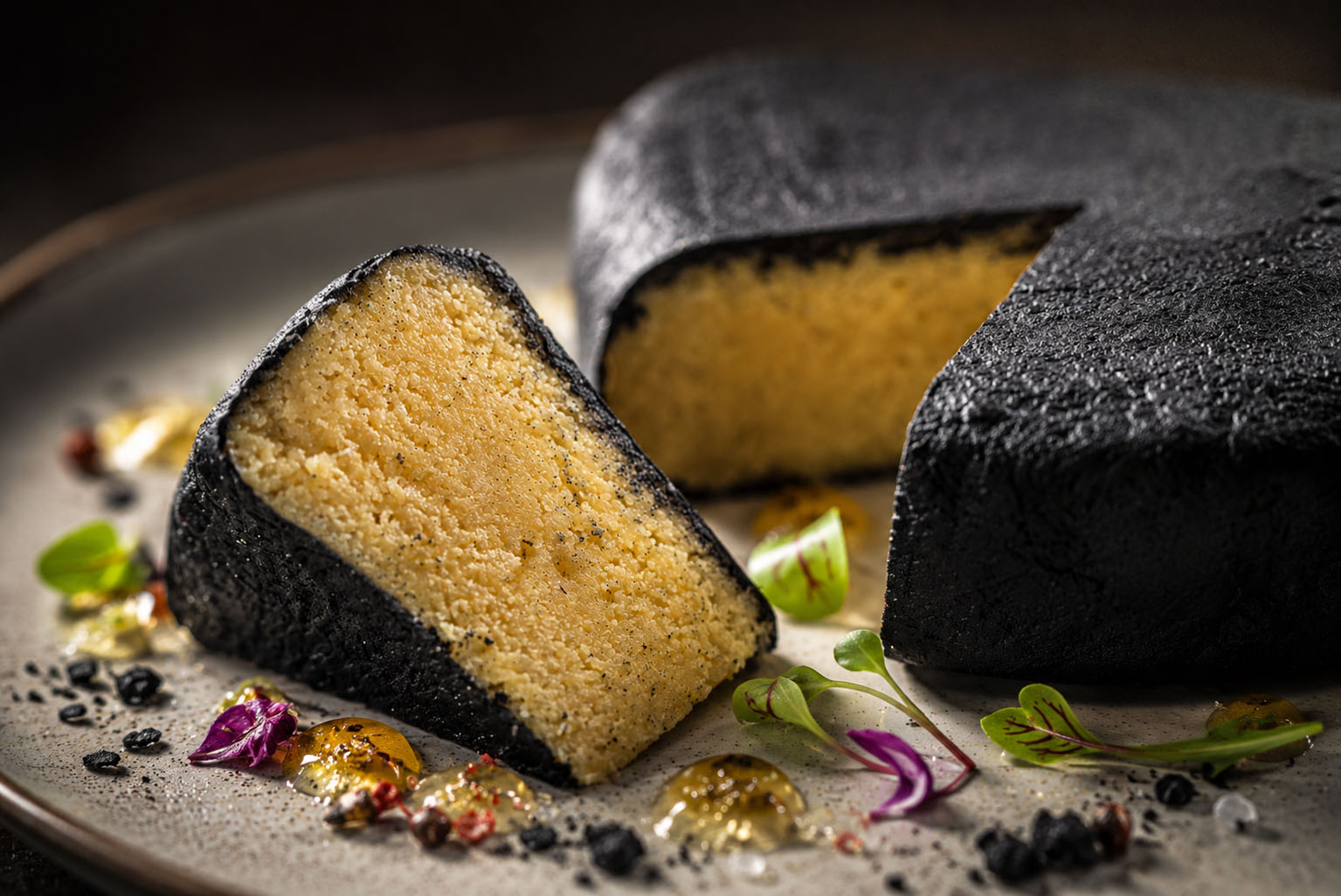 Slice of black cake with yellow interior on a plate, garnished with herbs and edible decorations, with the remaining cake in the background.