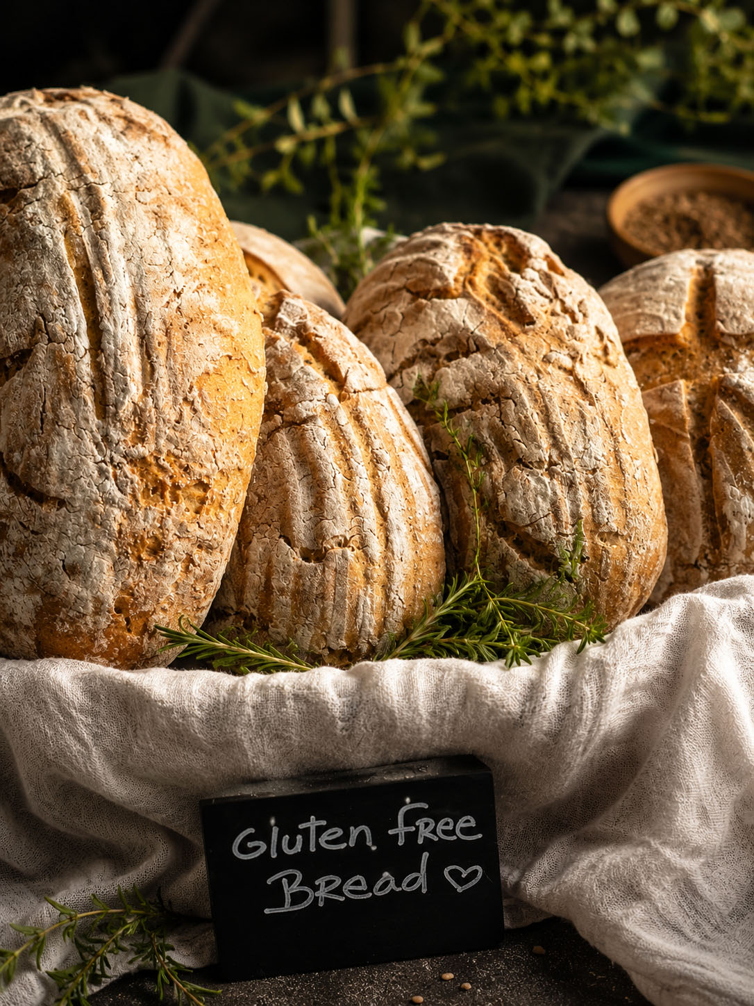 Four loaves of bread on a cloth with a sign reading 'Gluten free Bread' and greenery in the background.