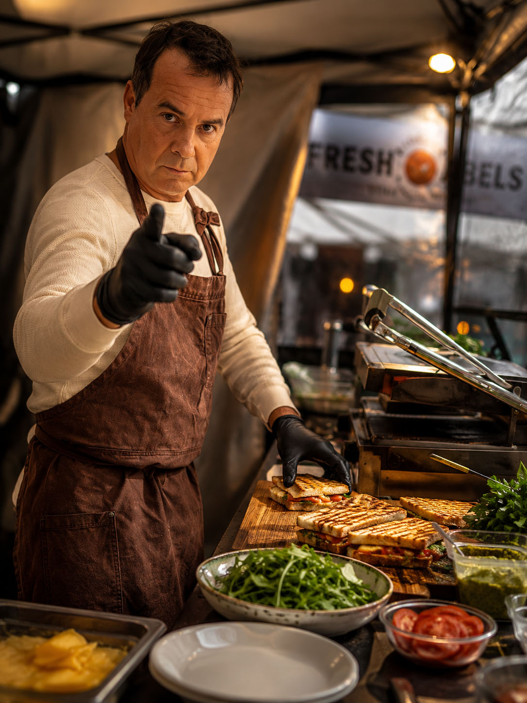 Man wearing apron and gloves preparing food at a stall with fresh ingredients, looking at camera.