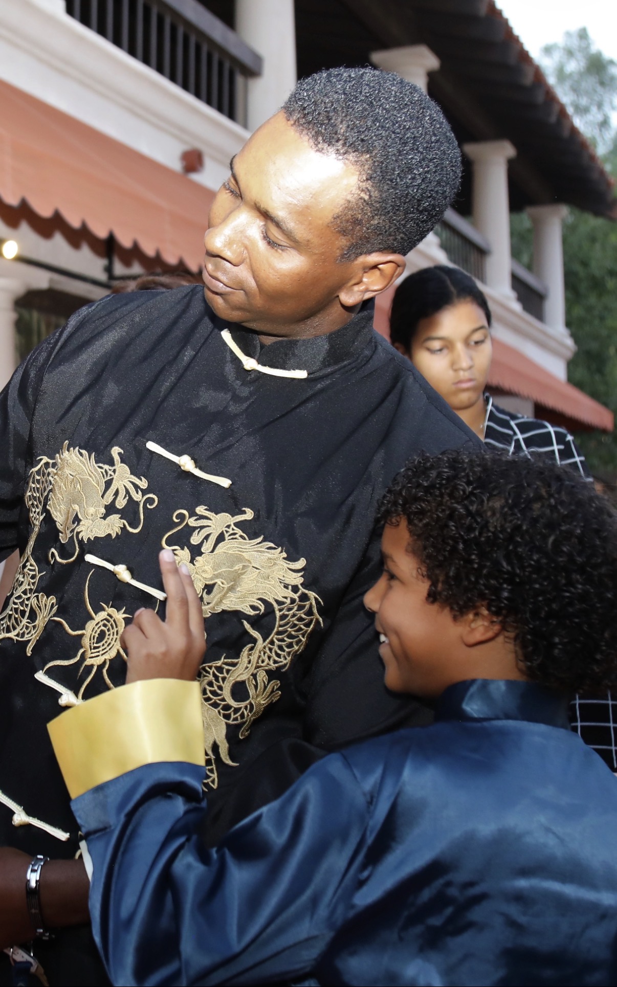 Two people outdoors, one in a black and gold embroidered outfit, the other in a blue jacket, smiling and interacting.