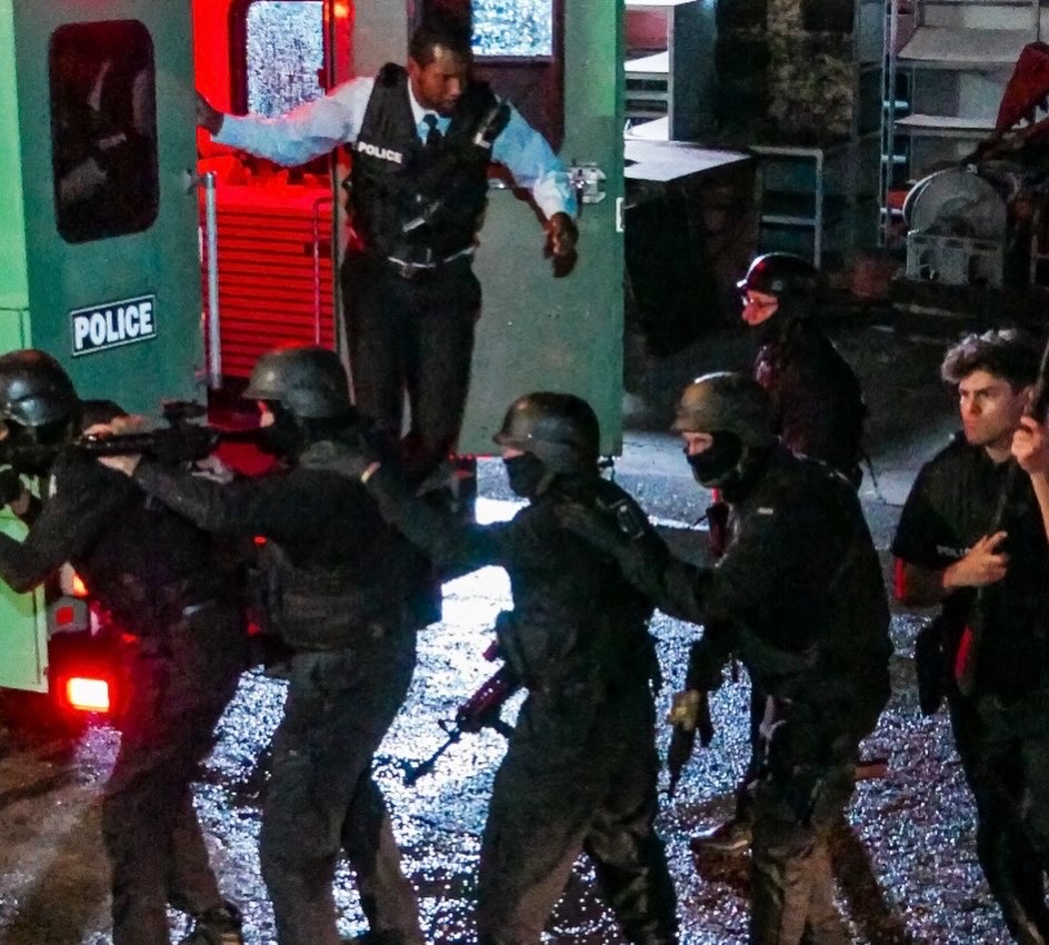 Group of police officers in black uniforms and masks, some holding guns, in a street scene at night.