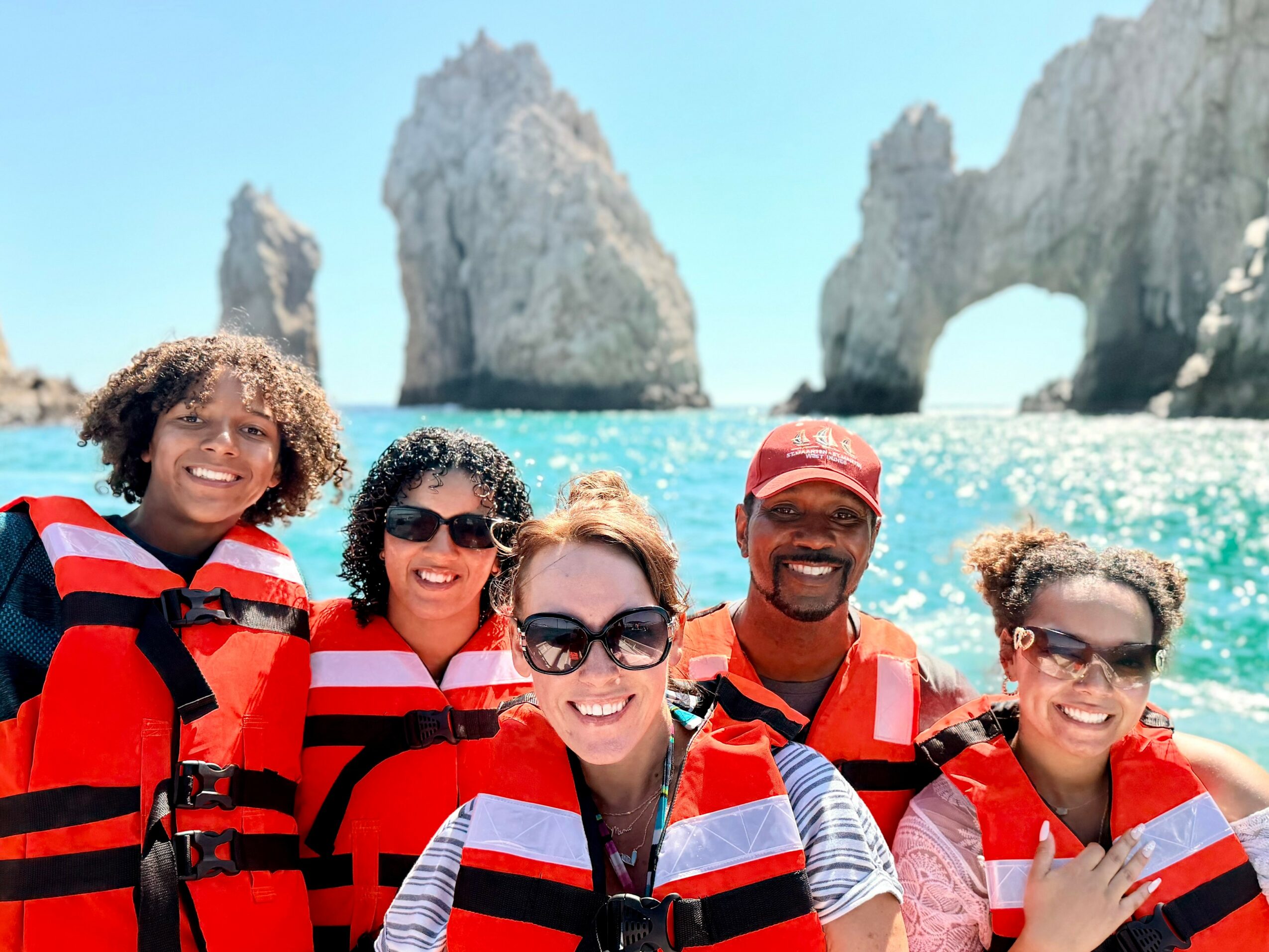 Group of five people in red life jackets smiling at the camera with ocean and rock formations in background.