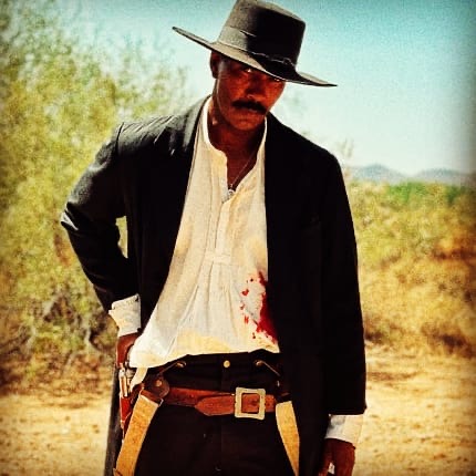 Man in cowboy hat and black jacket standing outdoors with desert background.