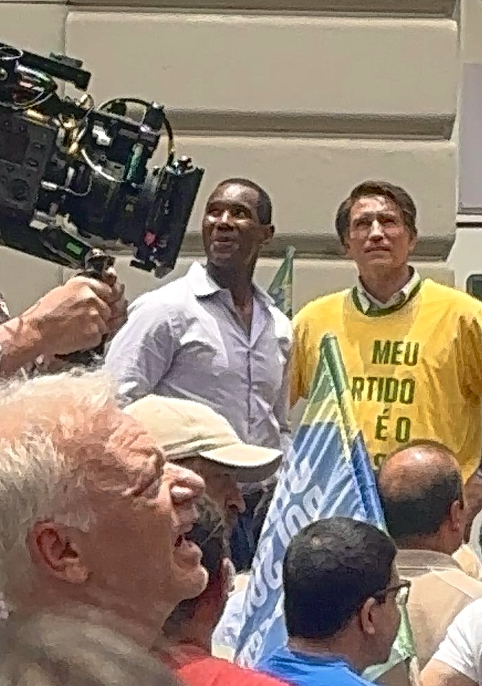 Two men standing outdoors, one in a white shirt and the other in a yellow shirt, with a crowd and camera equipment nearby.
