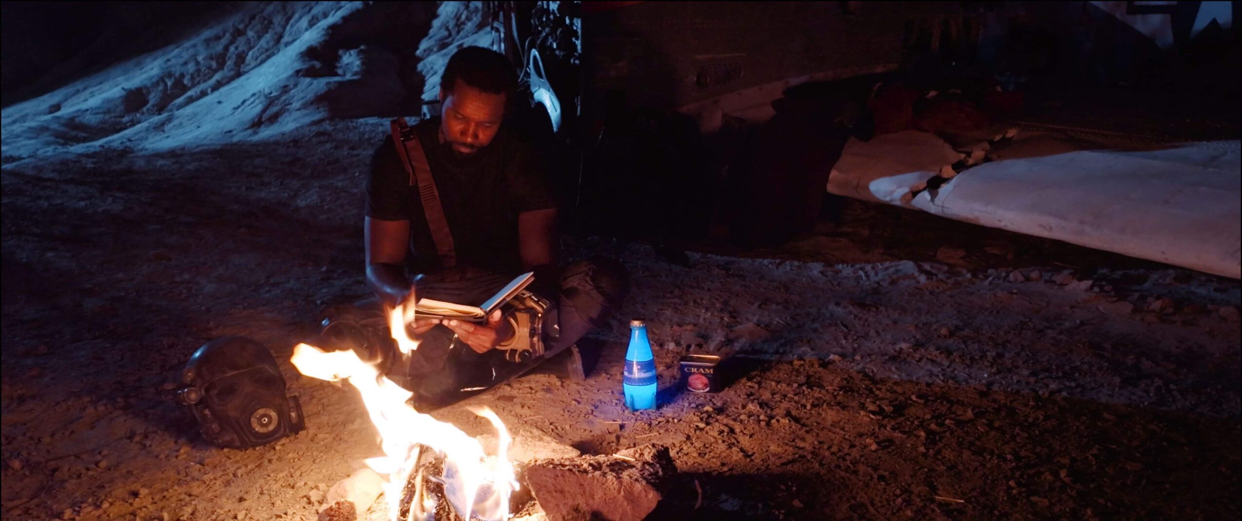 Person sitting near a campfire at night, reading a book, with a water bottle nearby, outdoors.