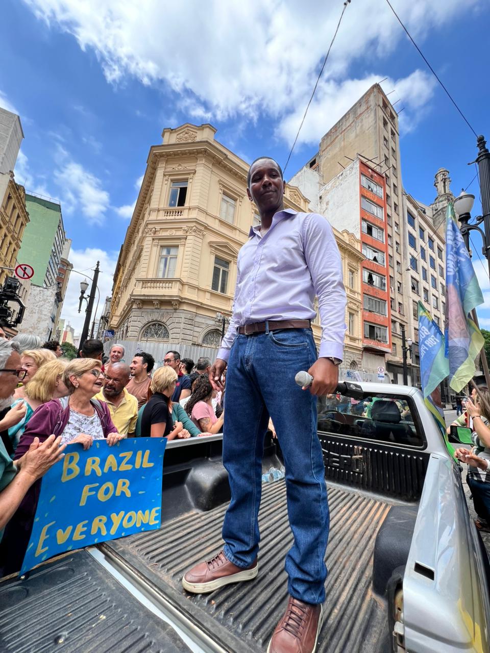 Man standing on vehicle with a crowd and buildings in the background during a street event.