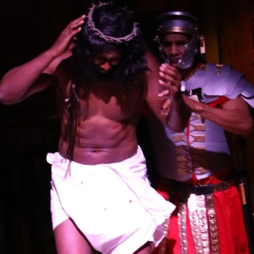 Person with long hair and a crown, shirtless, wrapped in a white cloth, assisted by a uniformed officer.