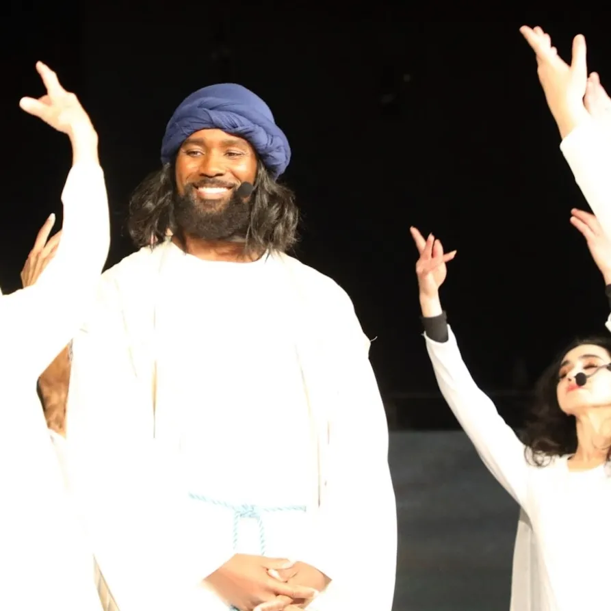 Person with long hair and beard wearing a blue head covering, smiling, with arms raised, on stage with others.