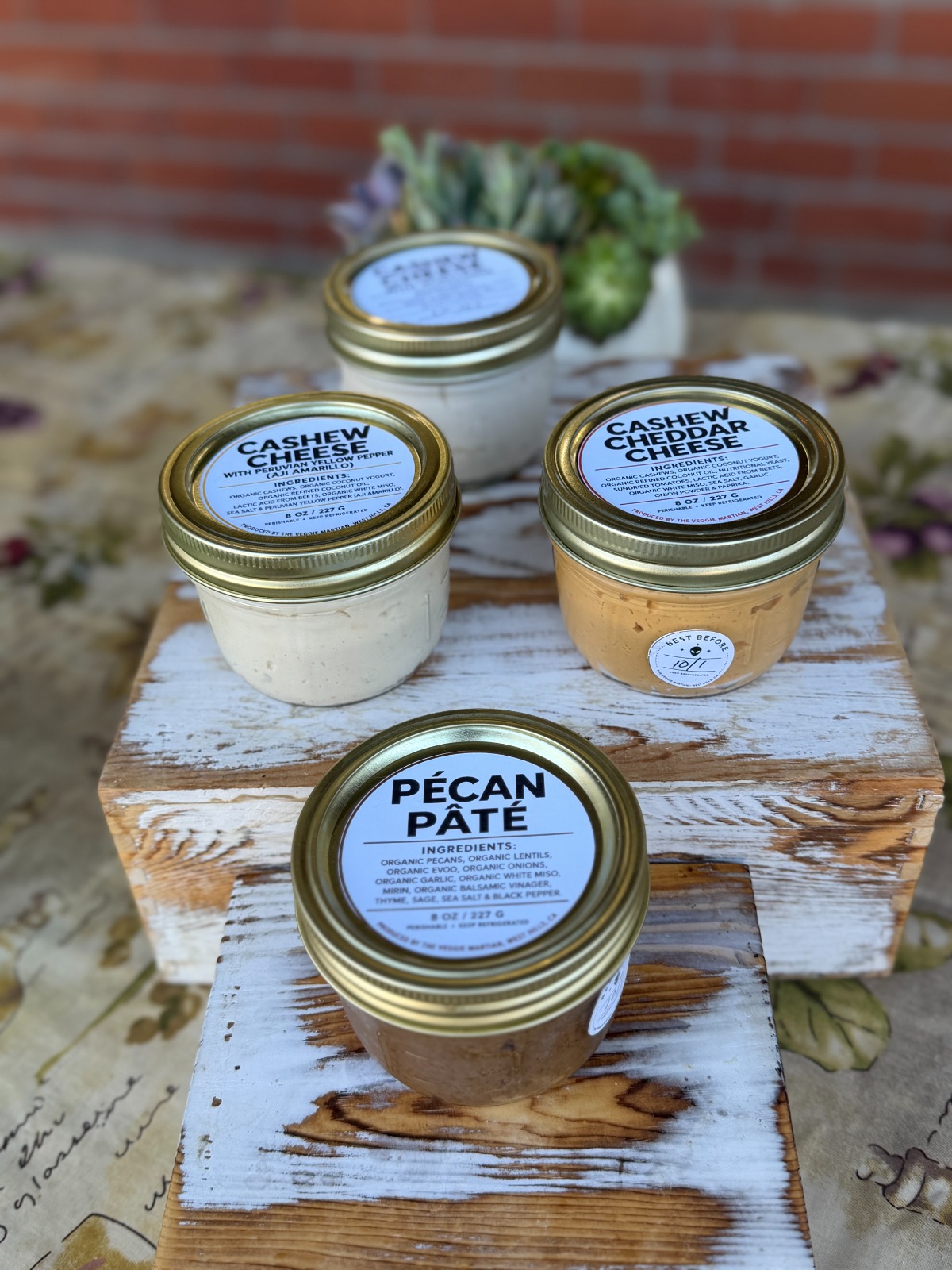 Four jars of spread placed on a rustic wooden surface with a brick wall and flowers in the background.
