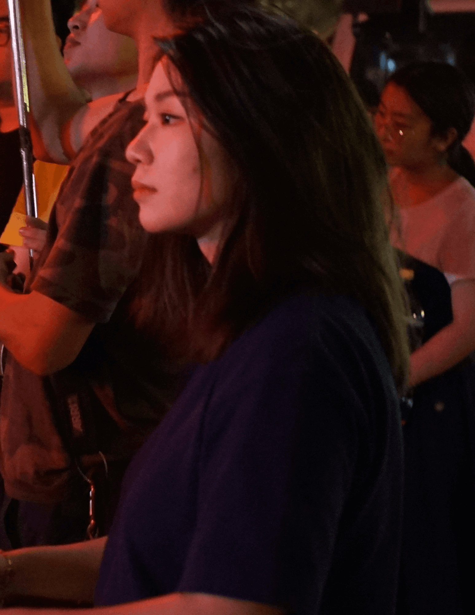 Side profile of a young woman with long dark hair, wearing a dark top, in a crowded indoor setting.
