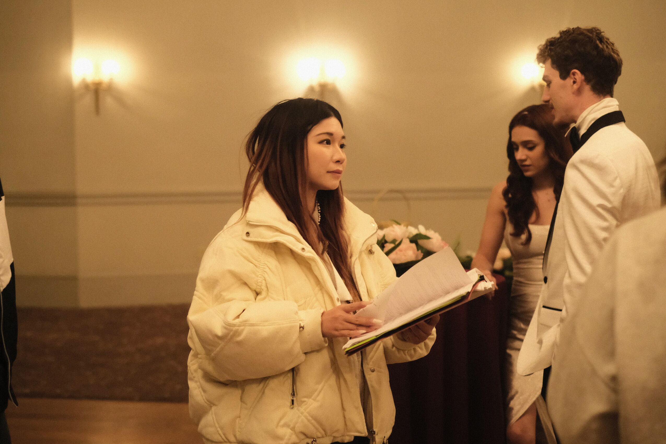 Two women and a man in formal attire at an indoor event, with warm lighting and a plain wall background.