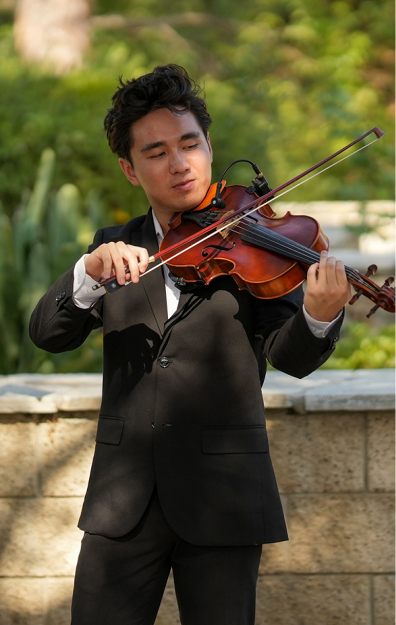 Young man in a black suit playing a violin outdoors with greenery in the background.