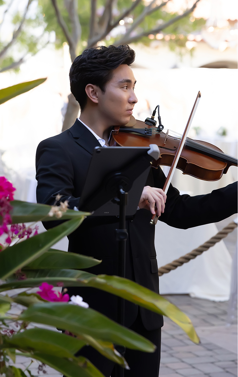 Young man playing violin outdoors, wearing a black suit, with greenery and flowers nearby.
