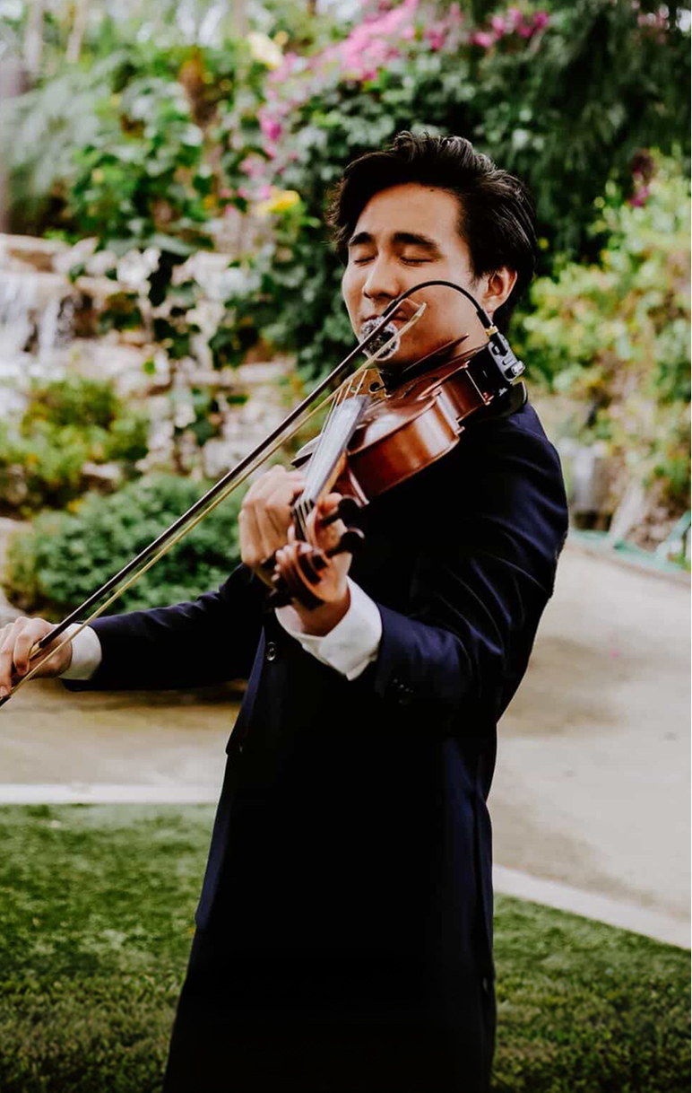 Person playing violin outdoors with greenery and flowers in background, eyes closed, wearing dark clothing.