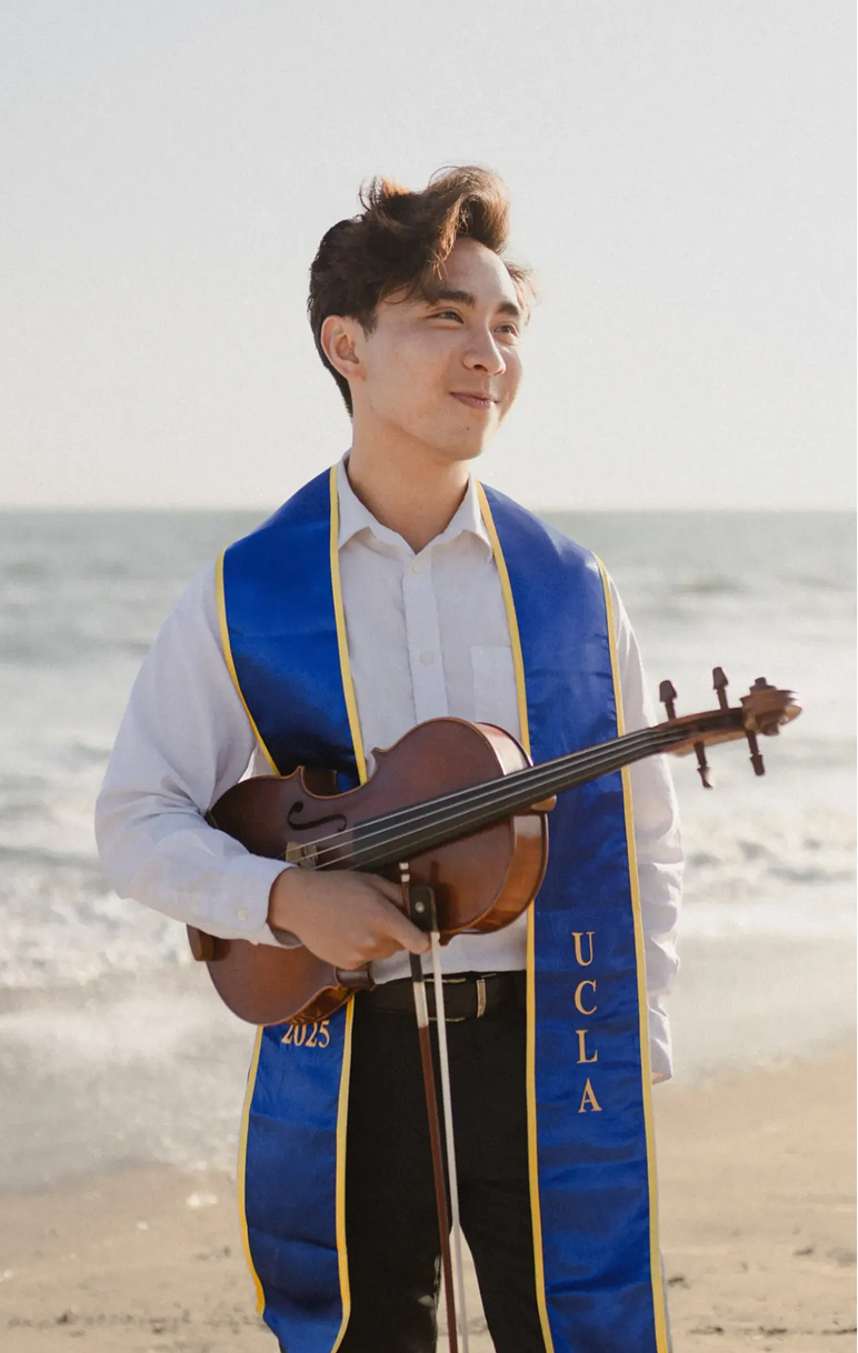 Young person standing on beach holding a small violin, wearing a white shirt and blue sash with 'UCLA' written on it.