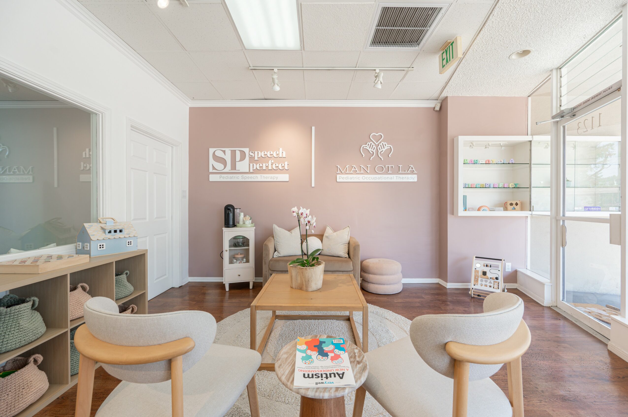 Waiting room with chairs, a coffee table, and a pink accent wall with signs and a crown, sunlight through windows.