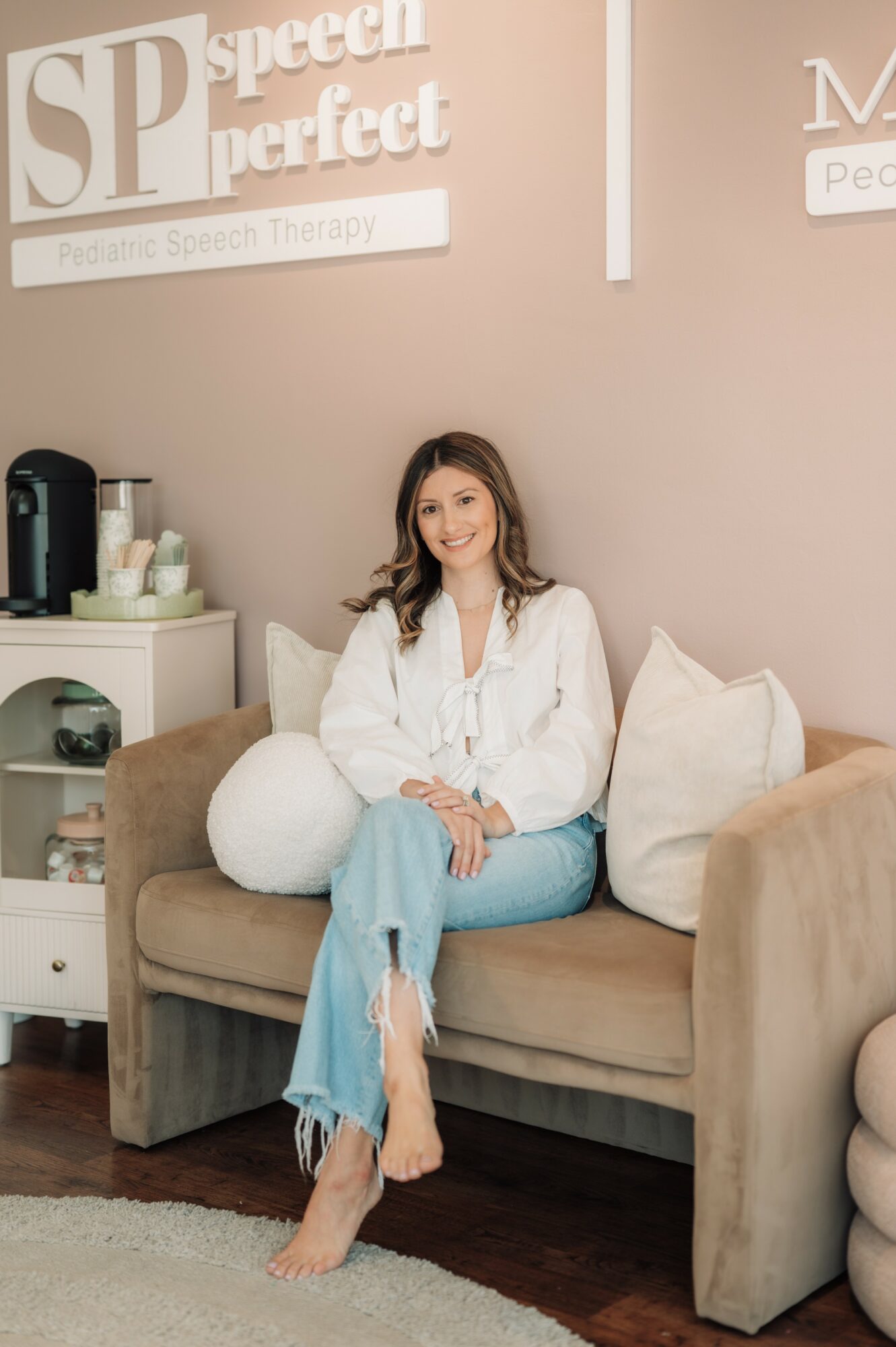Woman sitting on a beige sofa with pillows, smiling, in a room with speech therapy signs on the wall.