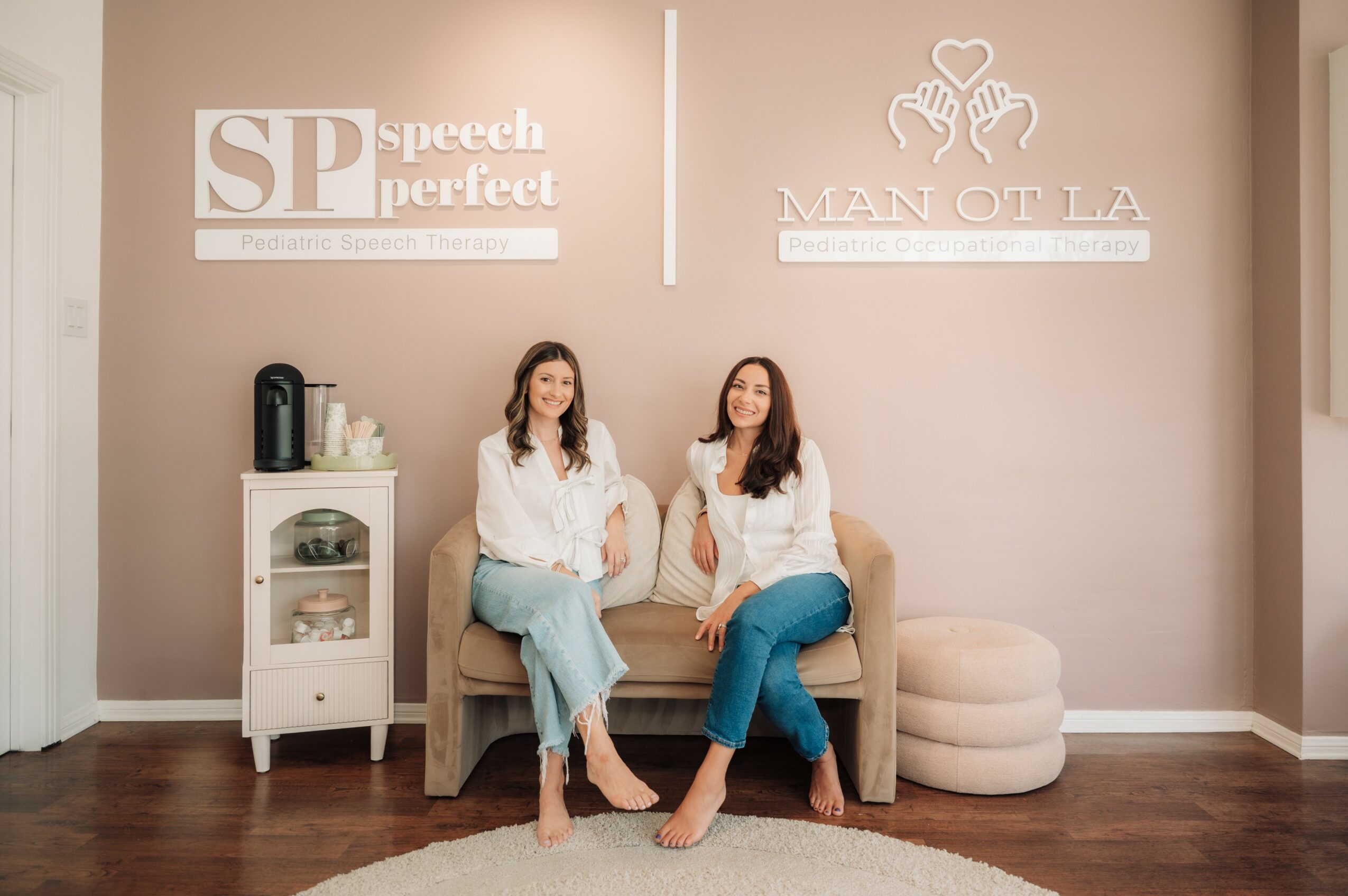 Two women sit on a beige sofa in a room with signs on the wall, one with long hair, both smiling, with a white cabinet nearby.