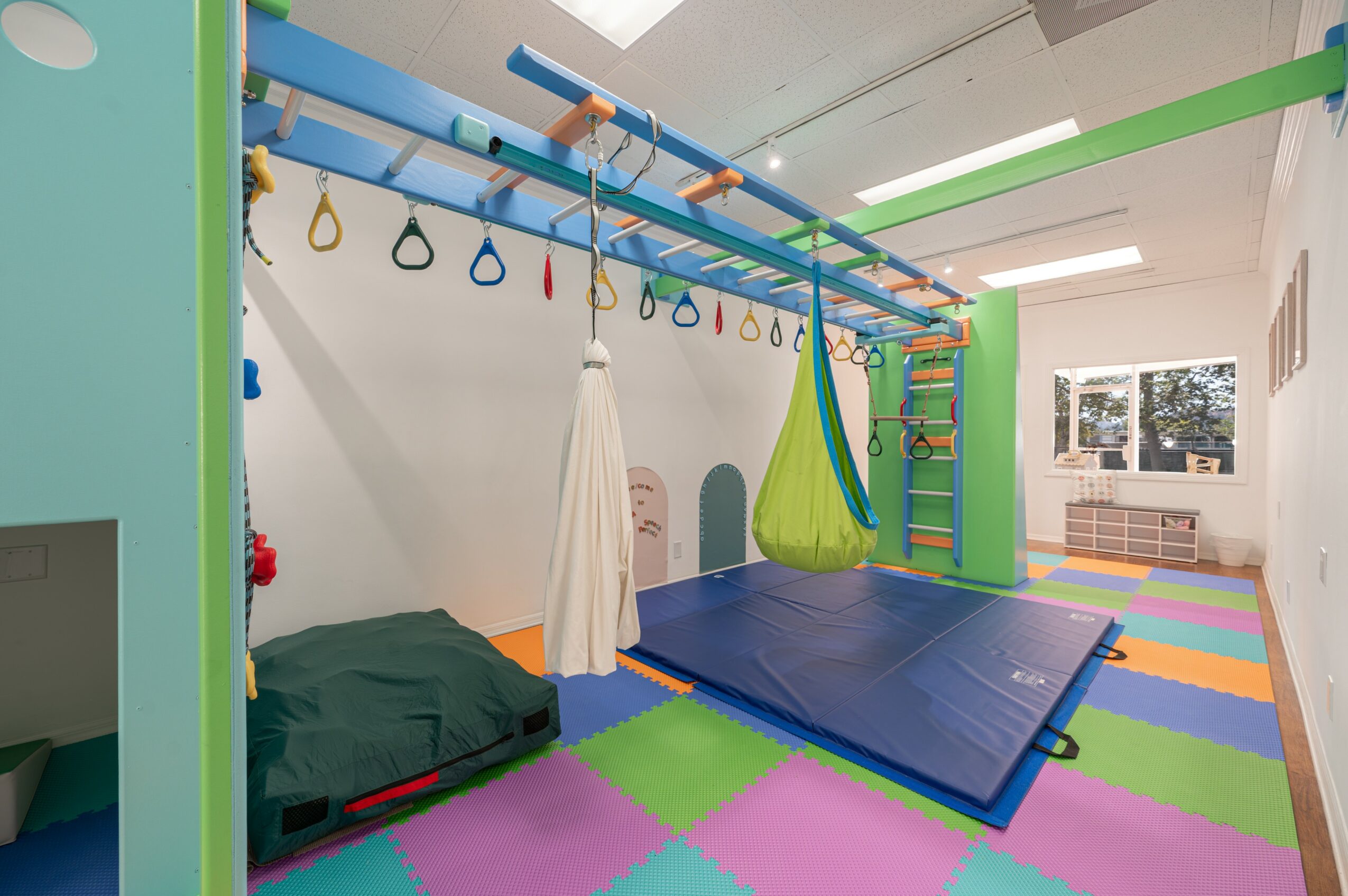 Indoor playroom with colorful foam mats, a bed, hanging rings, and climbing equipment, bright window light, and a white wall.