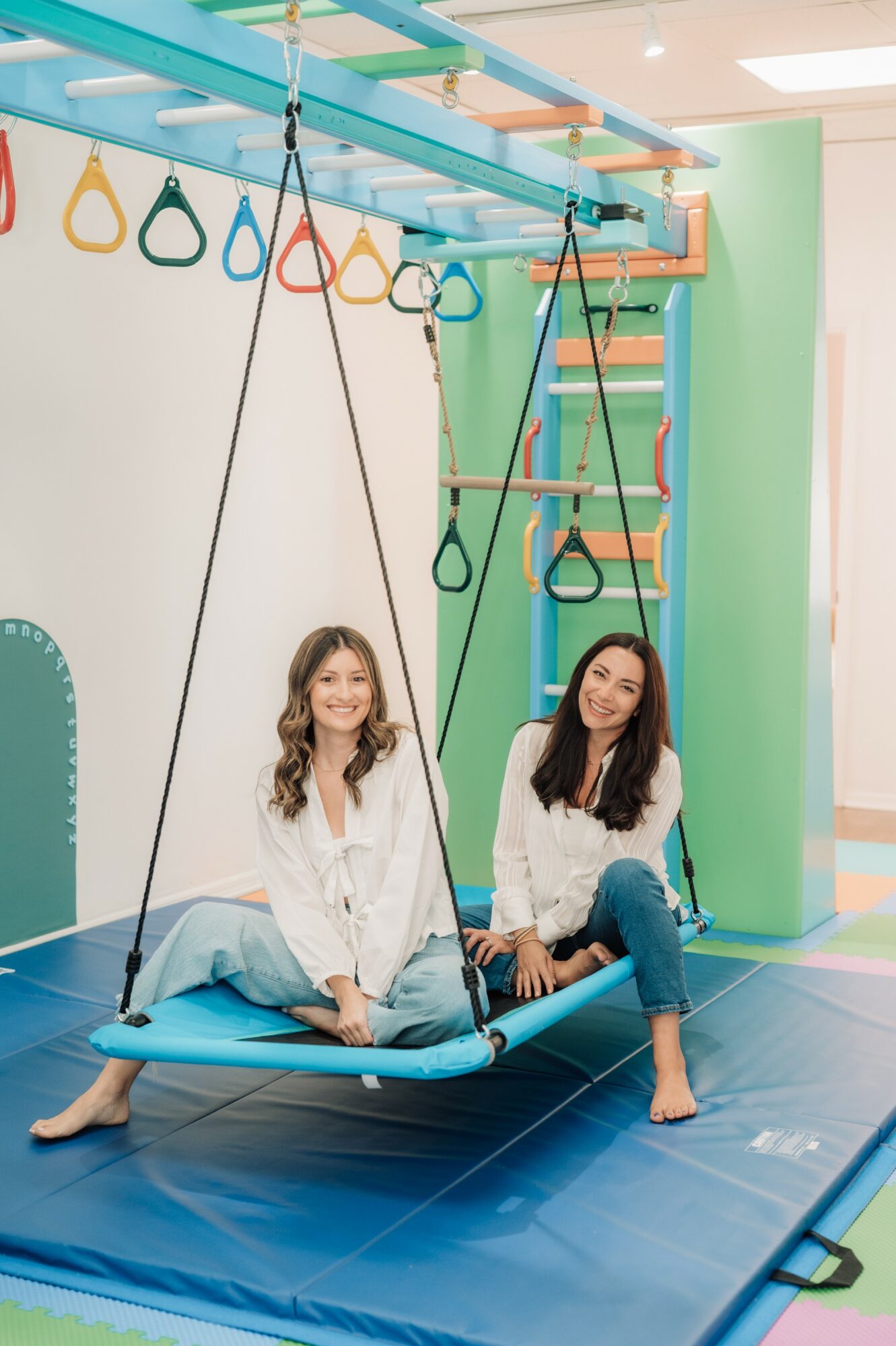 Two women sit on a blue swing in an indoor play area, smiling and looking at the camera.