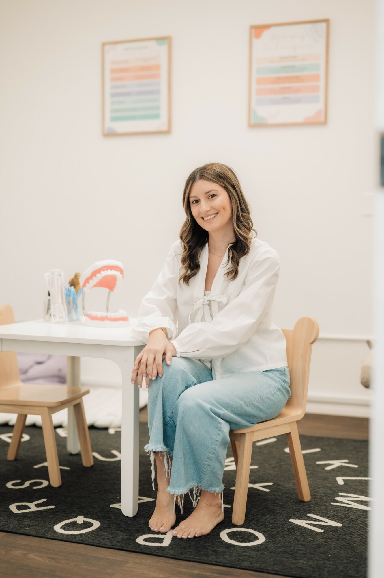 Smiling woman sitting on a chair at a table in a room with colorful framed artwork on the wall.