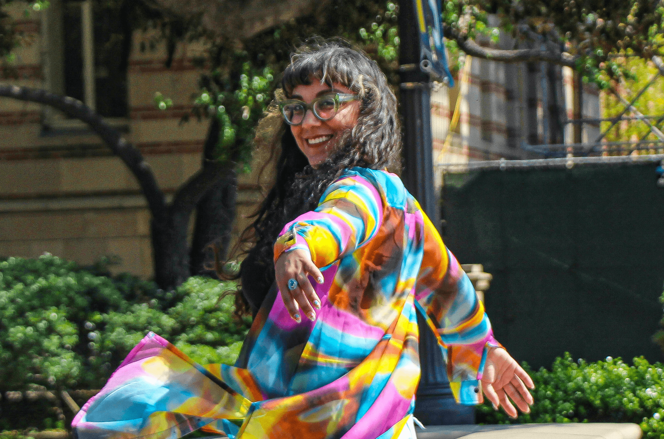 Smiling woman with glasses and long dark hair wearing a colorful, flowing dress outdoors with greenery and buildings in background.
